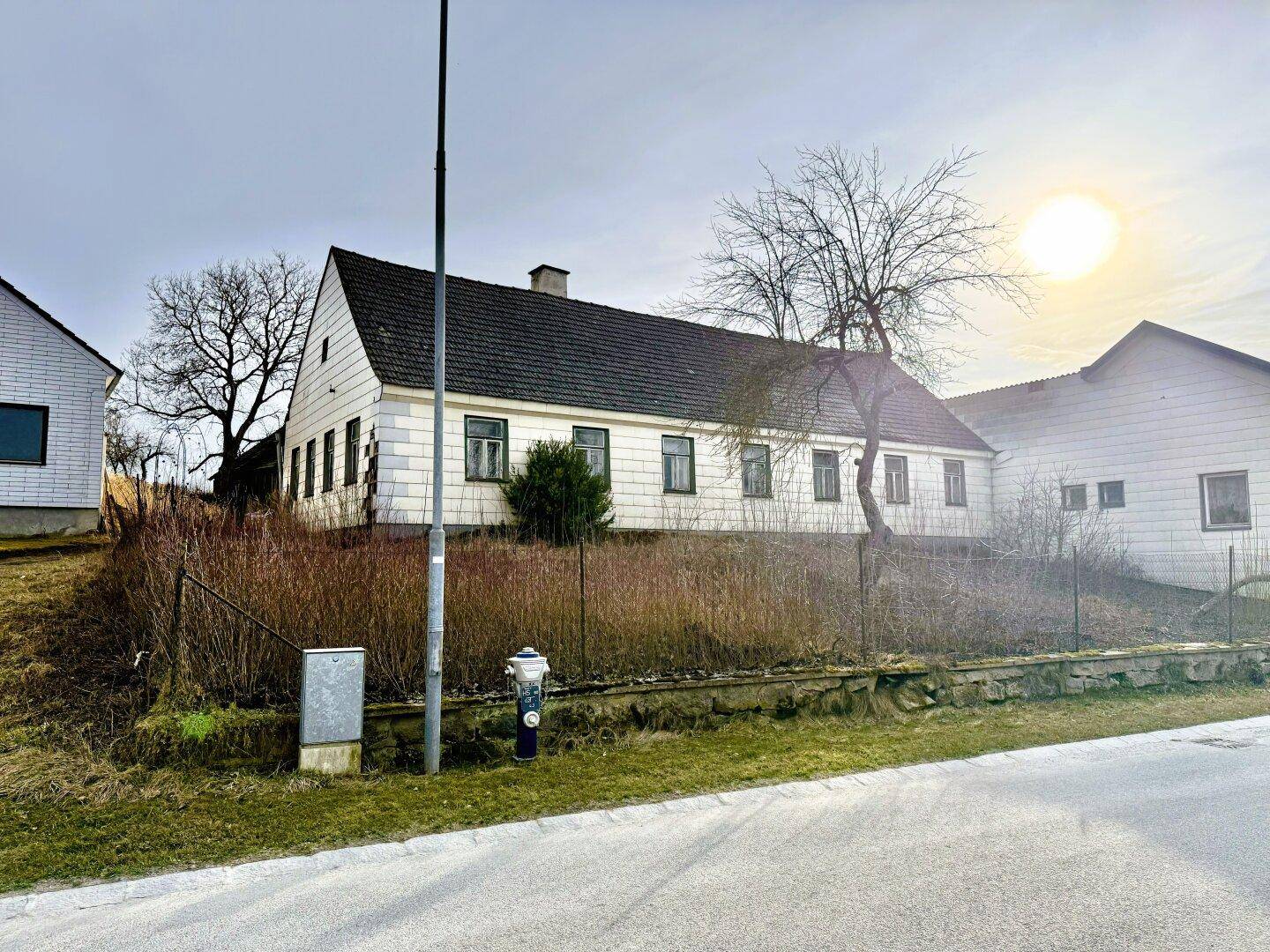 Altes, einfaches Haus mit Satteldach und einem ungepflegten Vorgarten an einer ländlichen Straße.