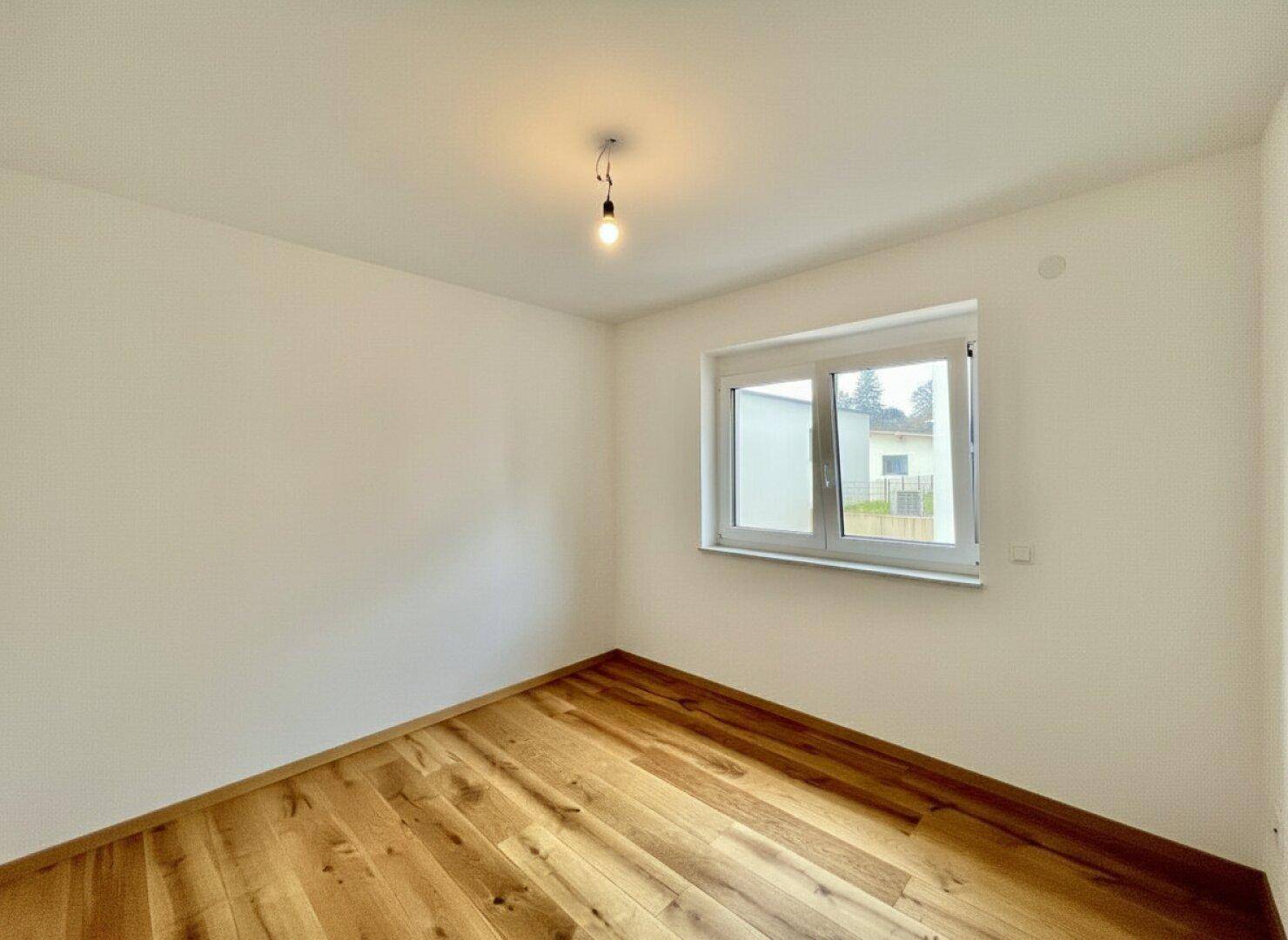 Freundliches, leeres Zimmer mit Holzboden und großem Fenster, ideal als Kinder- oder Gästezimmer.