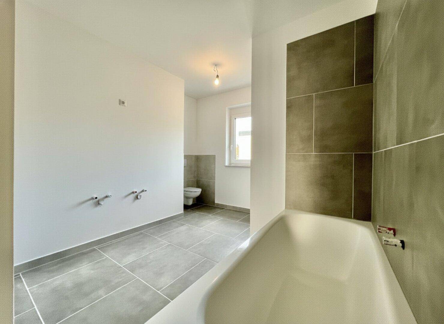 Modernes Badezimmer mit Badewanne, grauen Fliesen und Fenster für natürliche Belüftung.