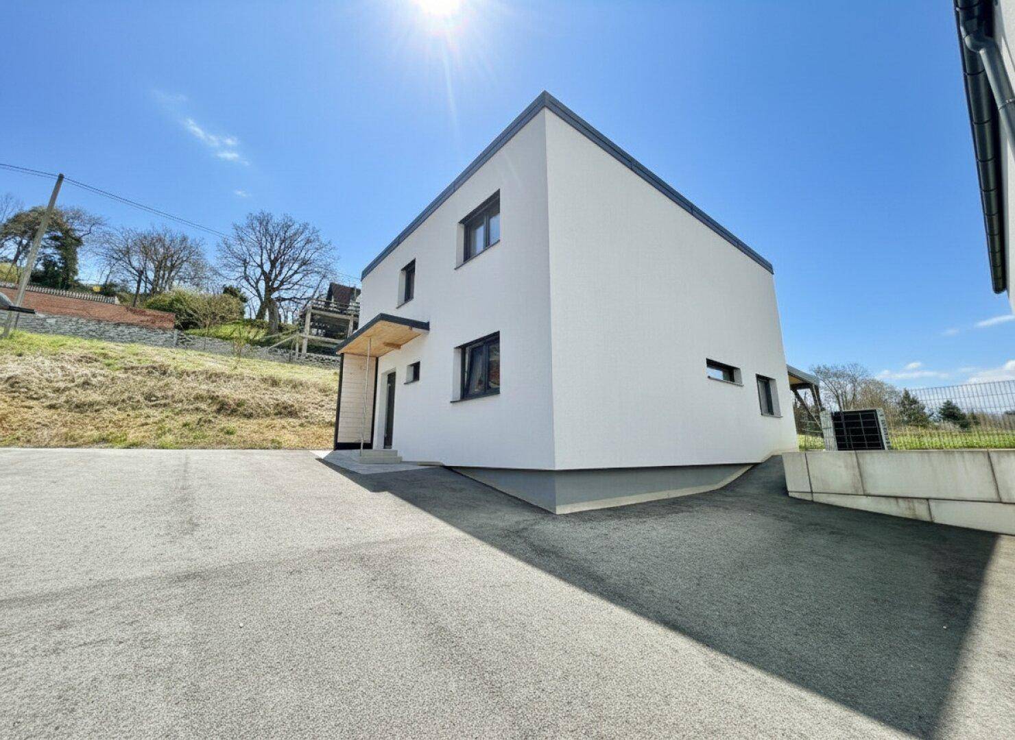 Modernes Einfamilienhaus mit weißer Fassade und Zufahrt, umgeben von Grünflächen unter blauem Himmel.