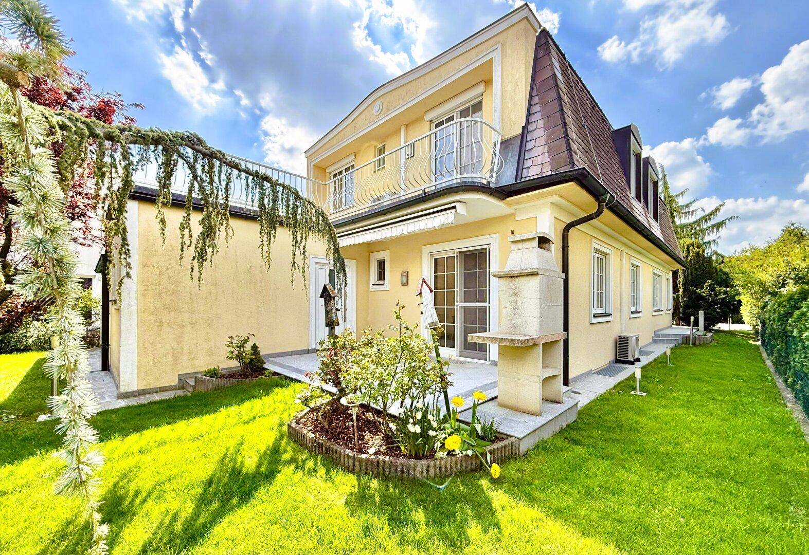 Rückansicht des Hauses mit gepflegtem Garten, Terrasse und Außenkamin unter blauem Himmel.