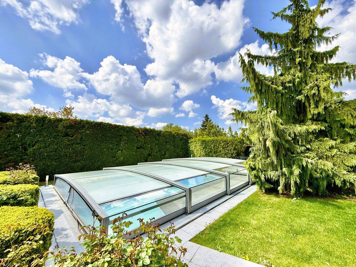 Überdachter Swimmingpool im Garten, umgeben von gepflegten Hecken und Rasenflächen unter blauem Himmel.