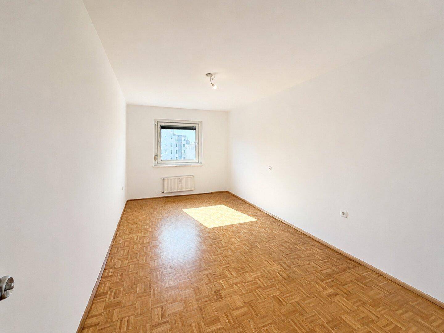 Leerer Raum mit Parkettboden und einem Fenster, das viel Tageslicht hereinlässt.