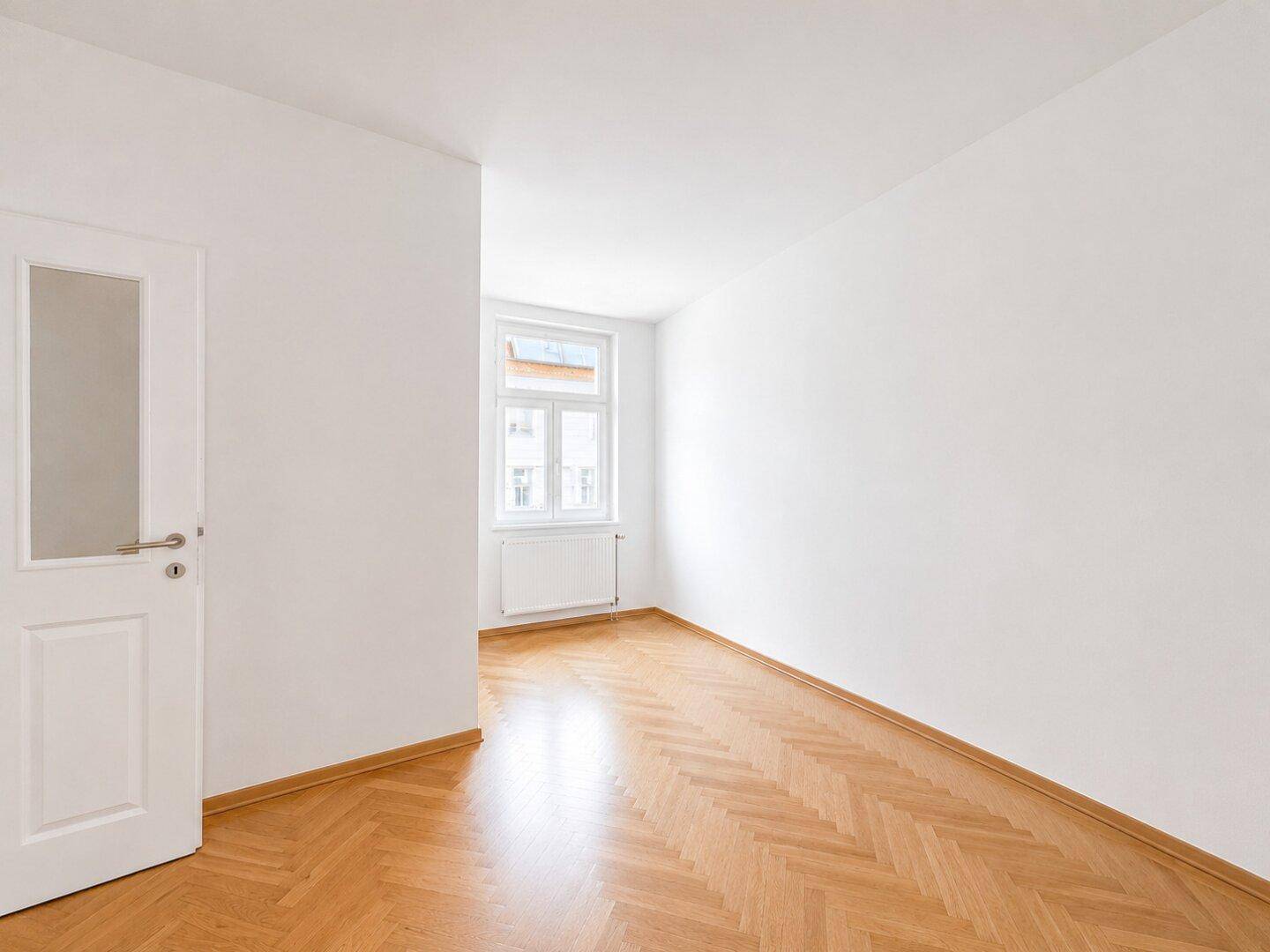 Helles, leeres Zimmer mit Fischgrätparkett und einem Fenster, ideal als Schlaf- oder Arbeitszimmer.