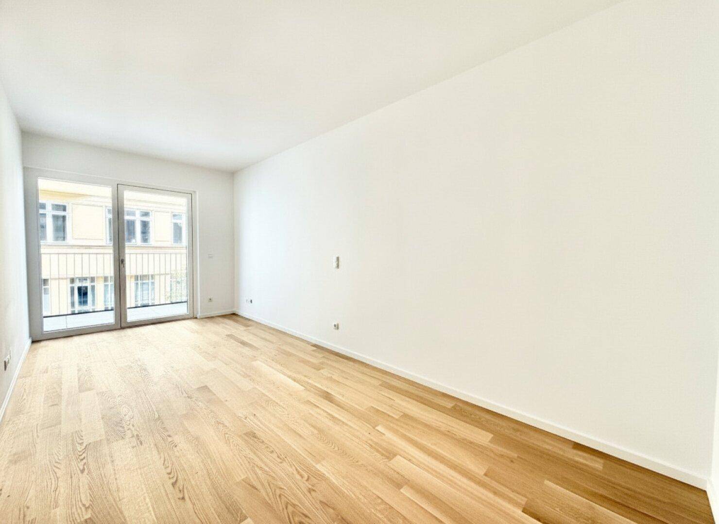 Leeres Schlafzimmer mit hellem Holzparkett und großer Schiebetür, die zum Balkon führt.
