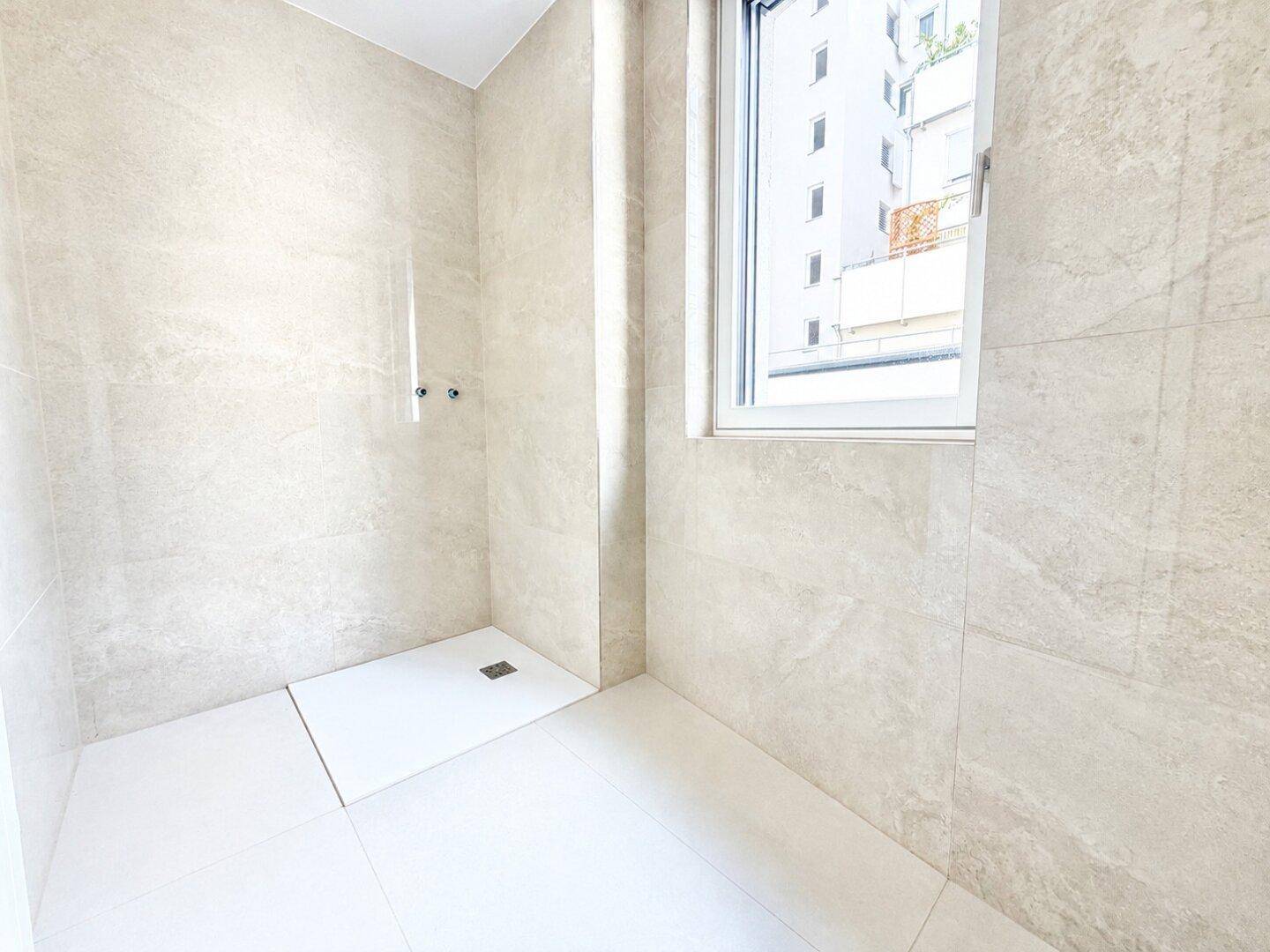 Modernes Badezimmer mit hellen Fliesen, ebenerdiger Dusche und Fenster für Tageslicht.