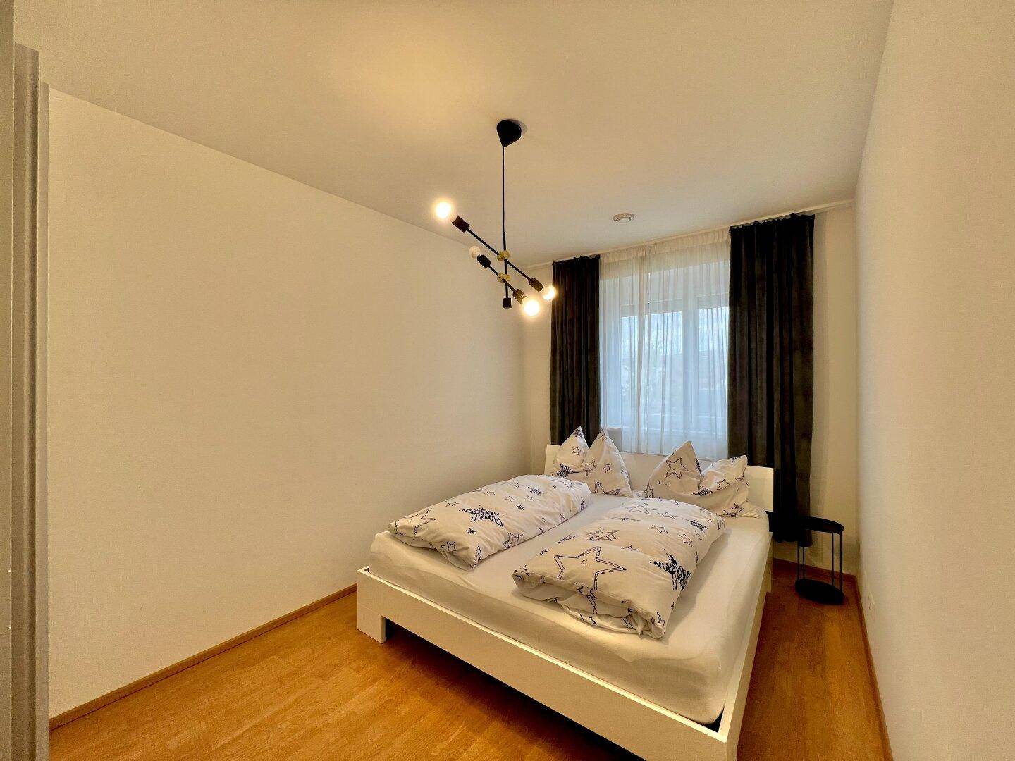 Gemütliches Schlafzimmer mit weißem Bett, dunklen Vorhängen und moderner Deckenleuchte.