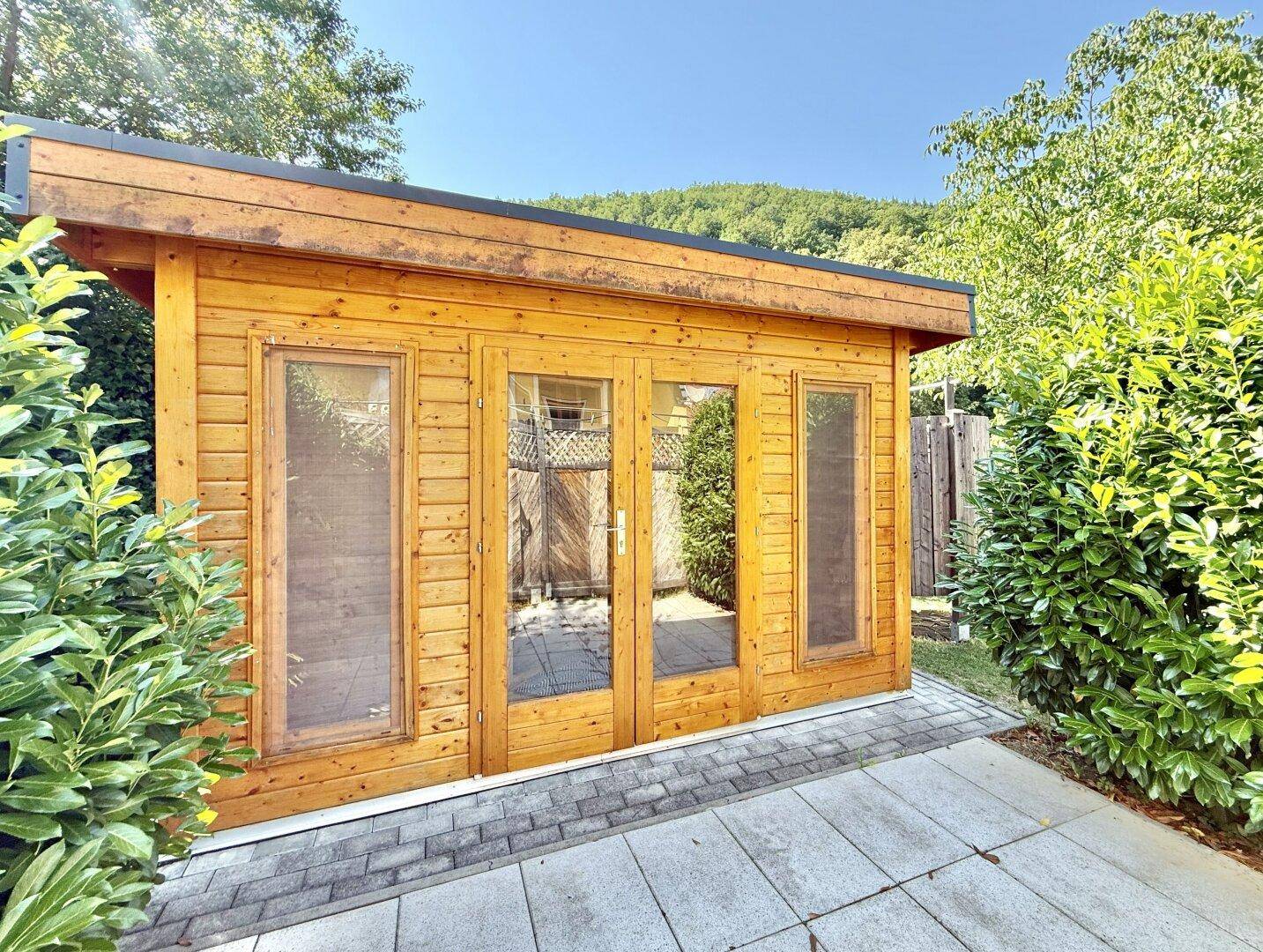 Moderne Gartenhütte aus Holz mit großen Glastüren, umgeben von grünen Büschen.
