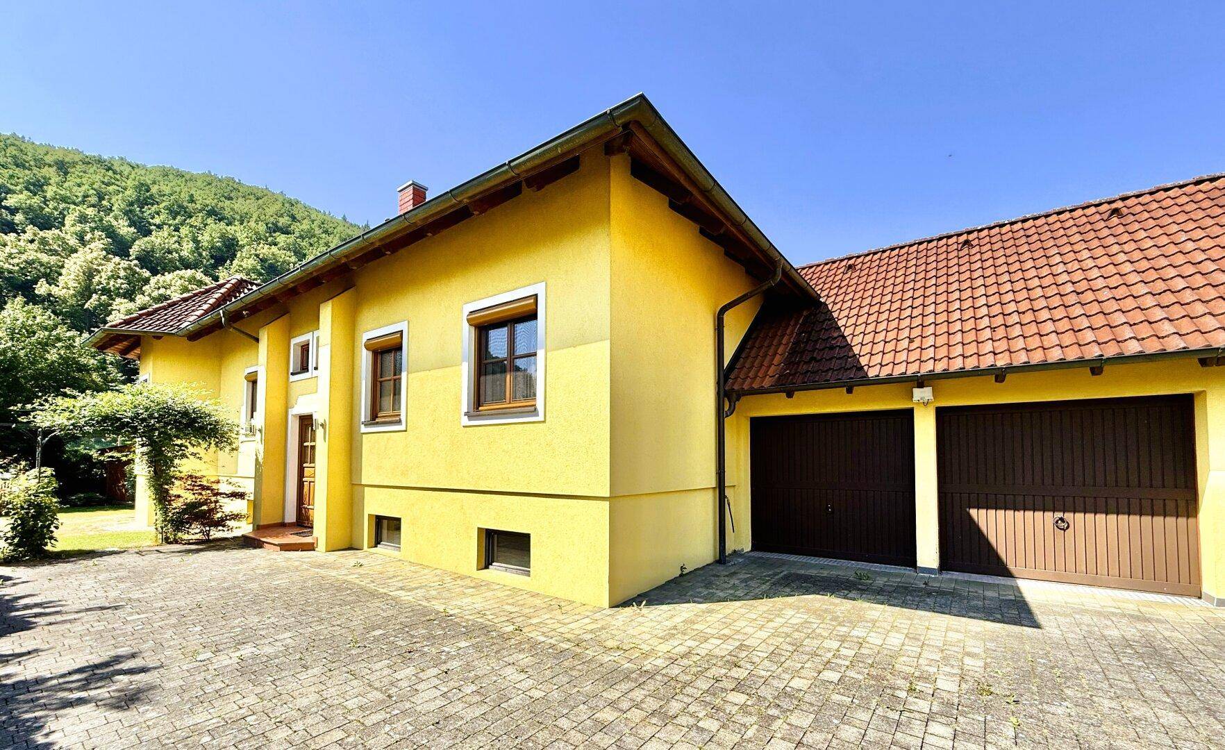Gelbes Haus mit zwei Garagen und gepflasterter Einfahrt, umgeben von grüner Landschaft.