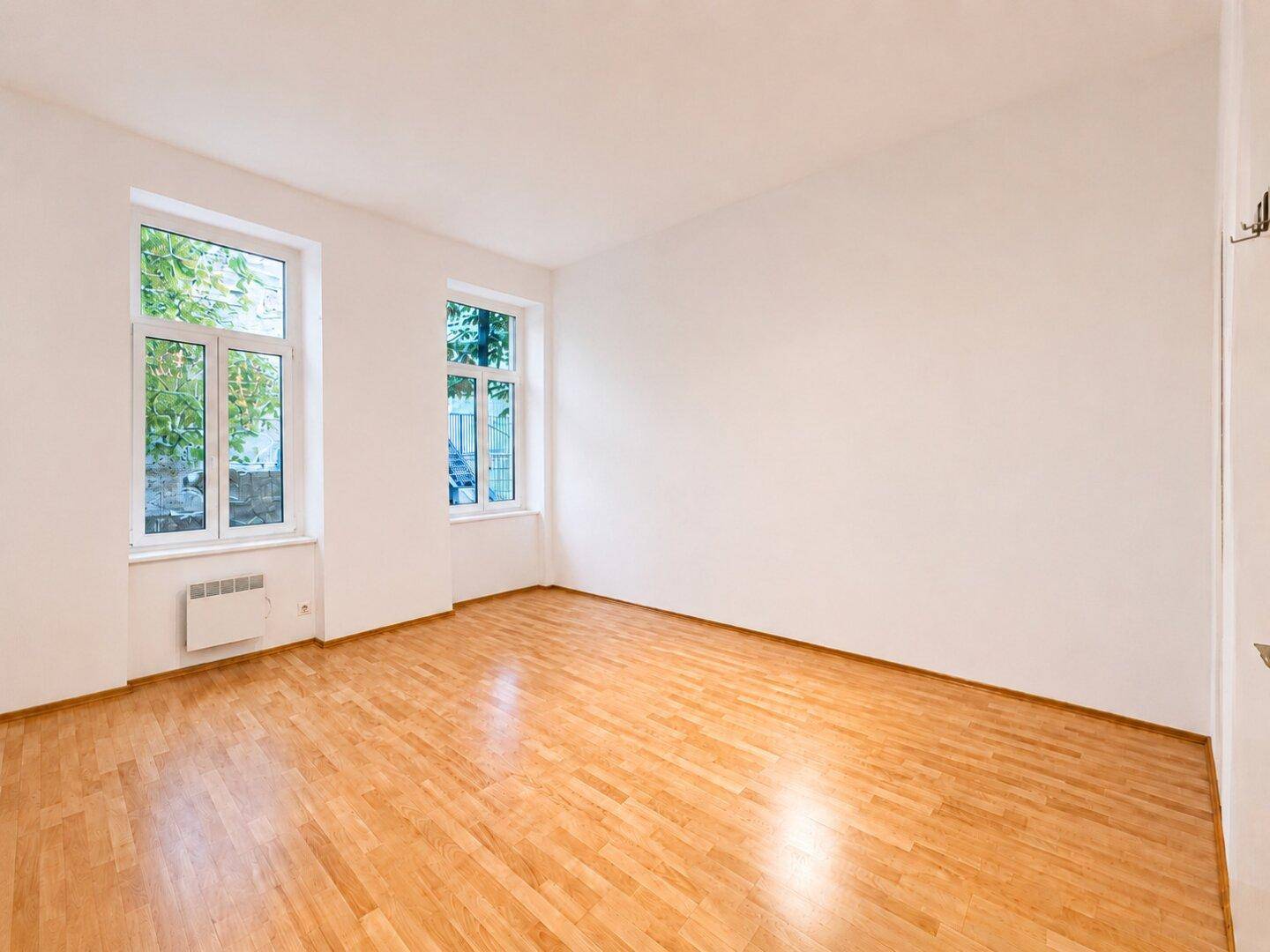 Geräumiges, leeres Zimmer mit hellem Holzboden und zwei großen Fenstern.