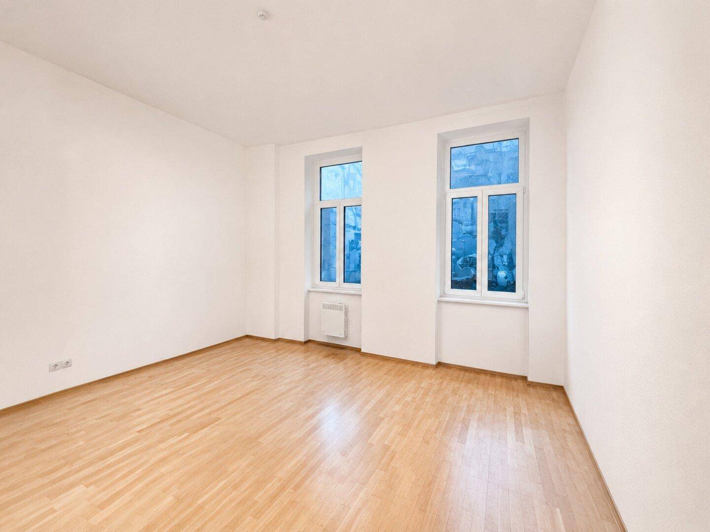 Leeres Zimmer mit Parkettboden und zwei Fenstern, bereit zur individuellen Gestaltung.