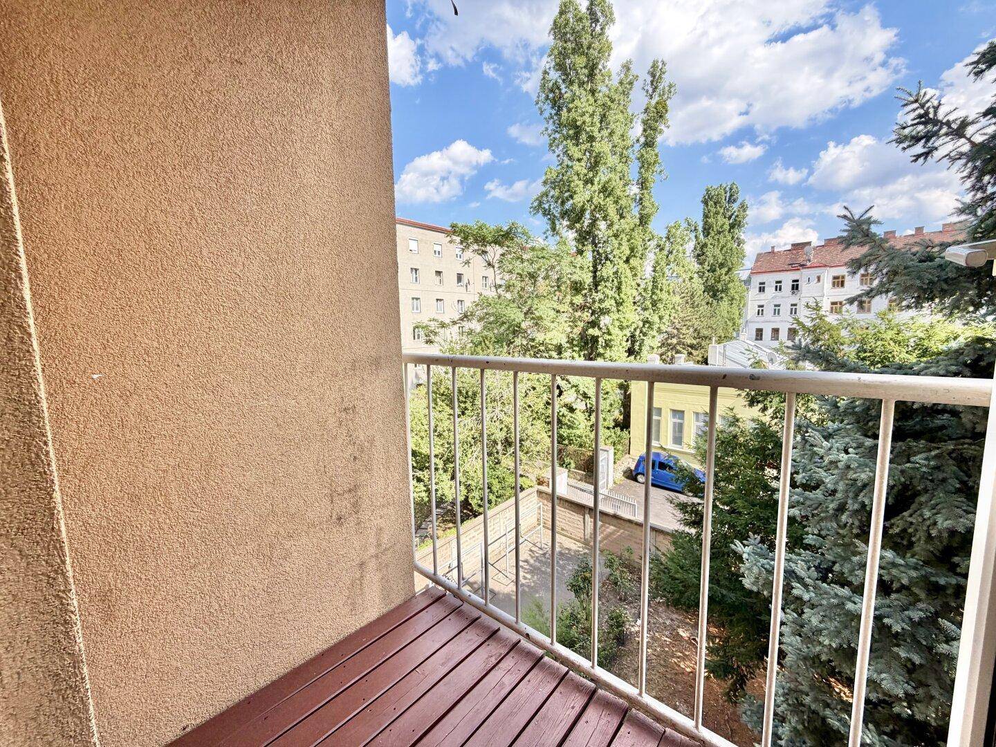 Kleiner Balkon mit Holzboden und Blick auf grüne Bäume und umliegende Gebäude.
