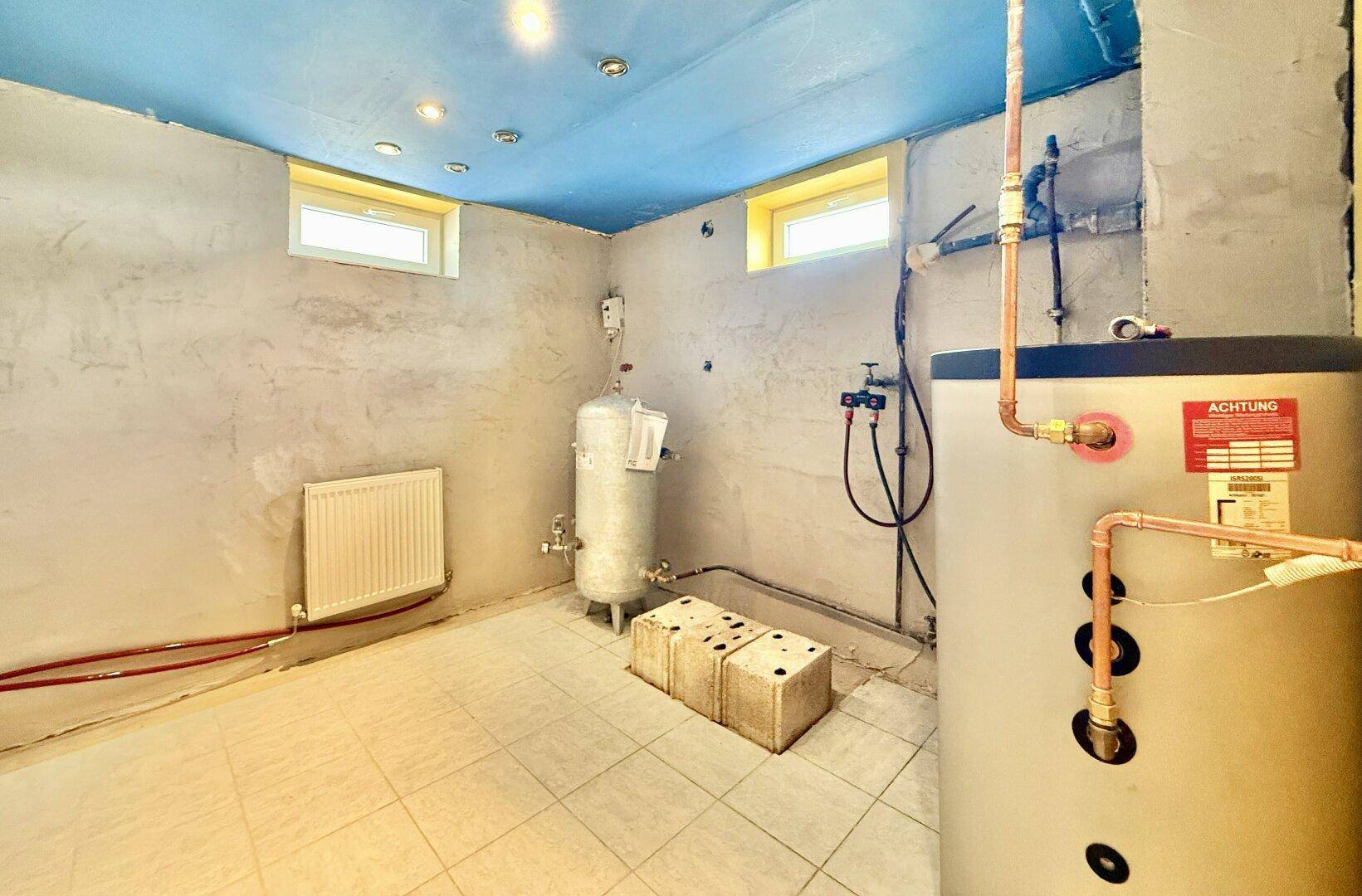 Hauswirtschaftsraum im Keller mit Warmwasserbereiter, sichtbaren Rohren und Fliesenboden.