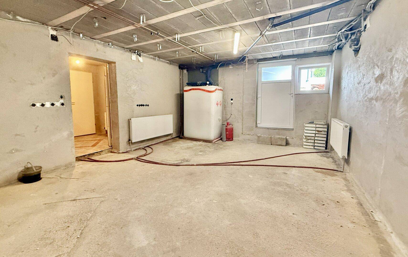 Großer Kellerraum mit Betonboden, Warmwasserbereiter und Zugang nach draußen.