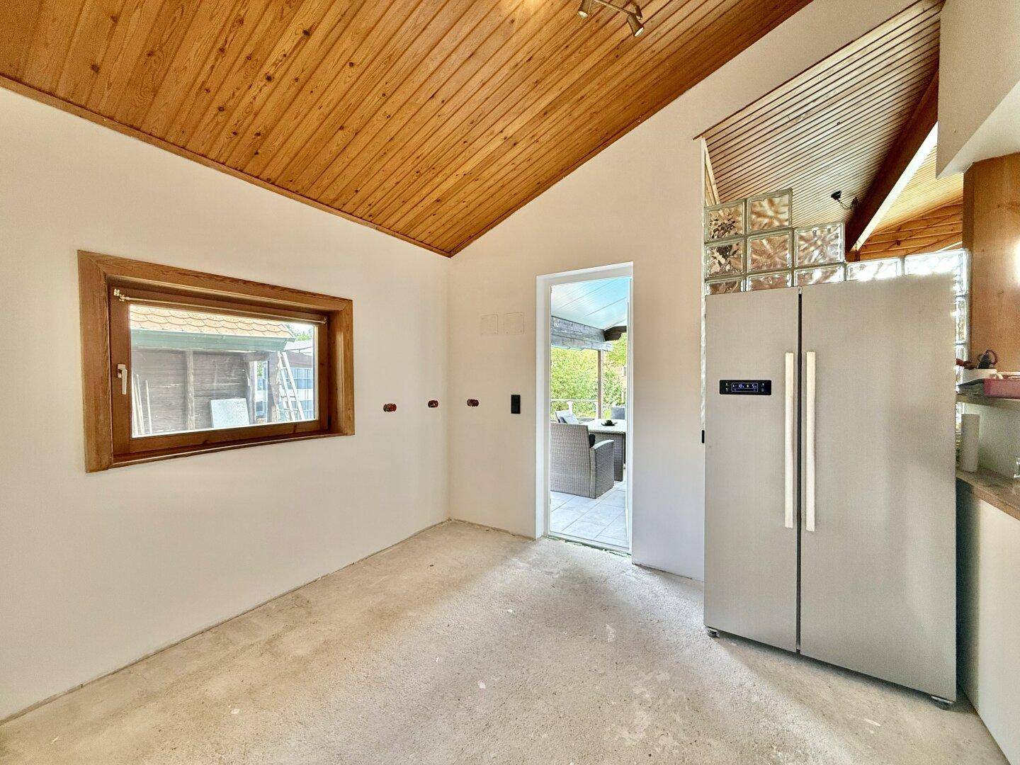 Leerer Küchenraum mit Holzdecke, großem Kühlschrank und Zugang zur Terrasse.