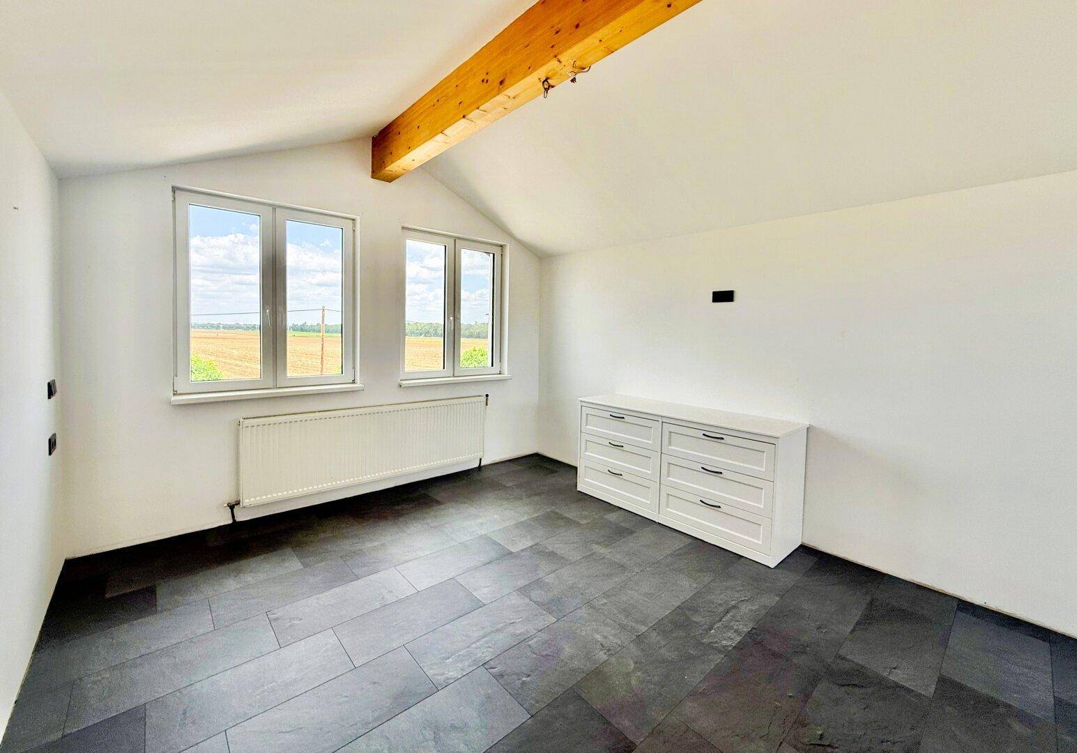 Leeres Schlafzimmer mit Holzbalken, großen Fenstern und weitem Blick auf die Landschaft.
