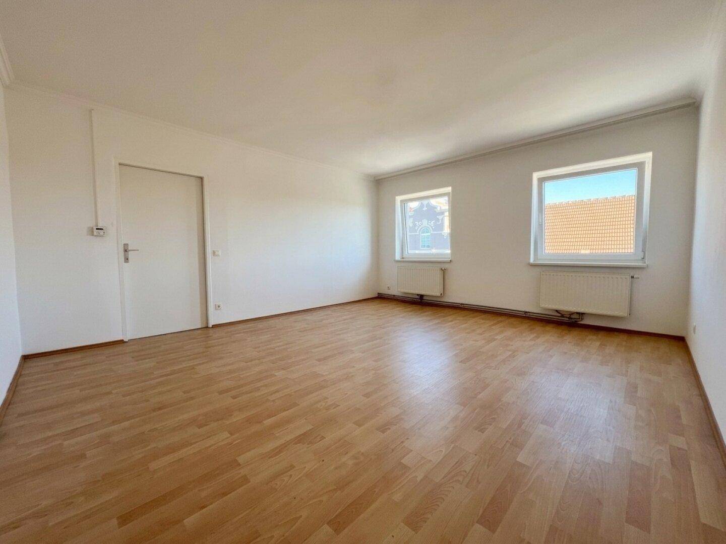 Geräumiges Zimmer mit Laminatboden, zwei Fenstern und weiß gestrichenen Wänden.