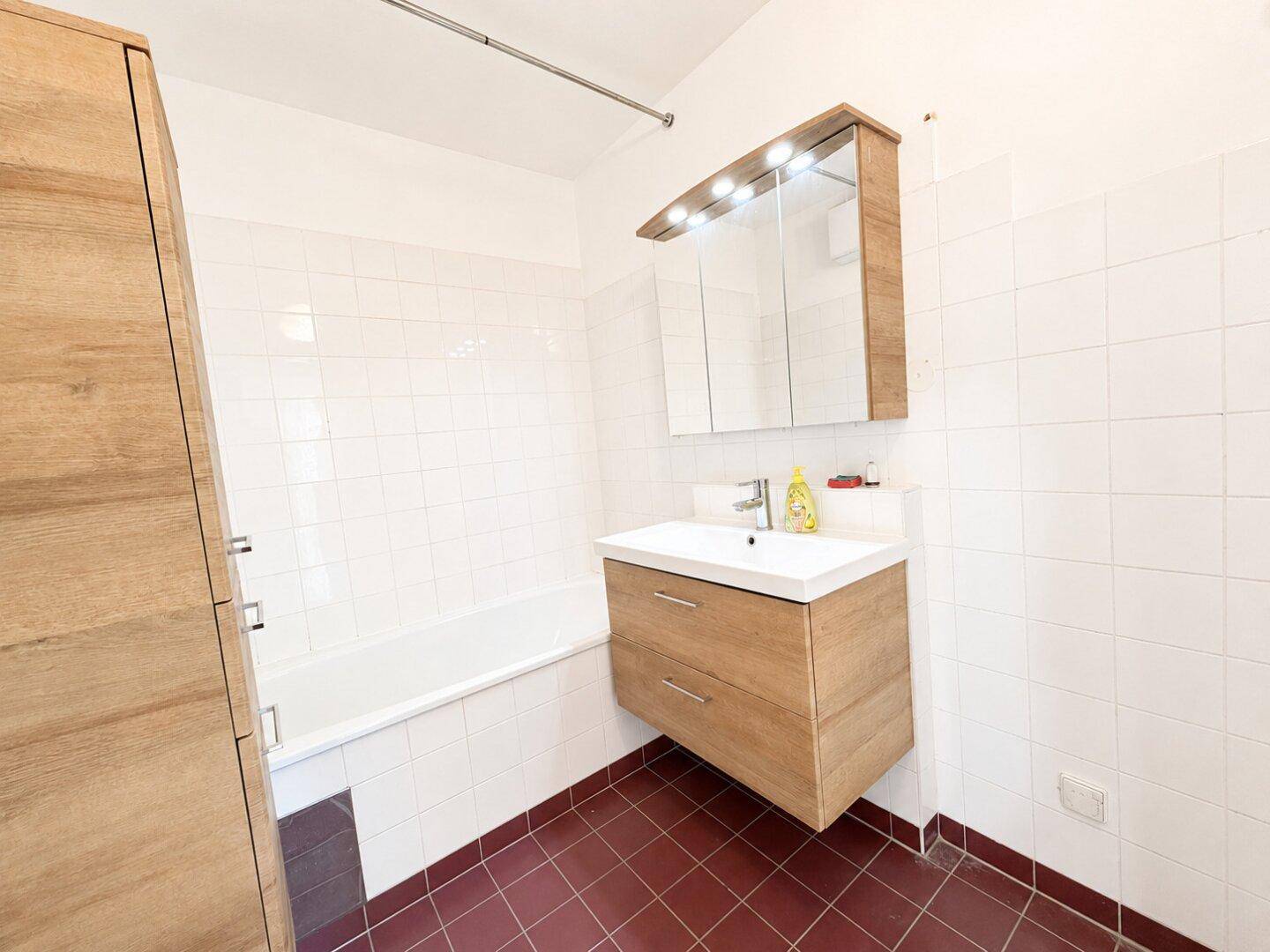 Badezimmer mit Badewanne, Einzelwaschbecken und Holzschrank, ausgestattet mit weißen und roten Fliesen.
