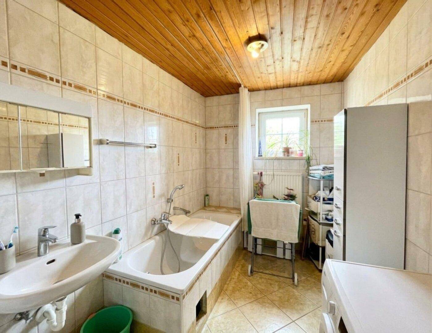 Badezimmer mit Badewanne, Waschbecken, hellen Fliesen und Holzdecke.