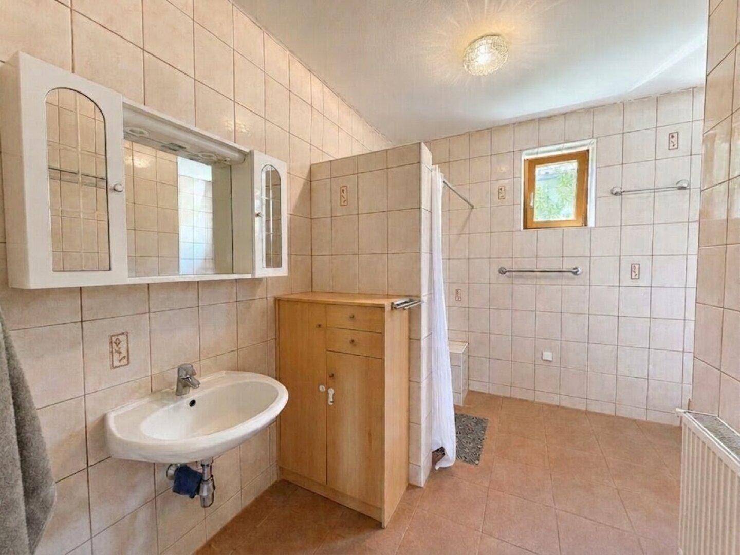 Helles Badezimmer mit beigen Fliesen, Dusche, Waschbecken und Fenster.