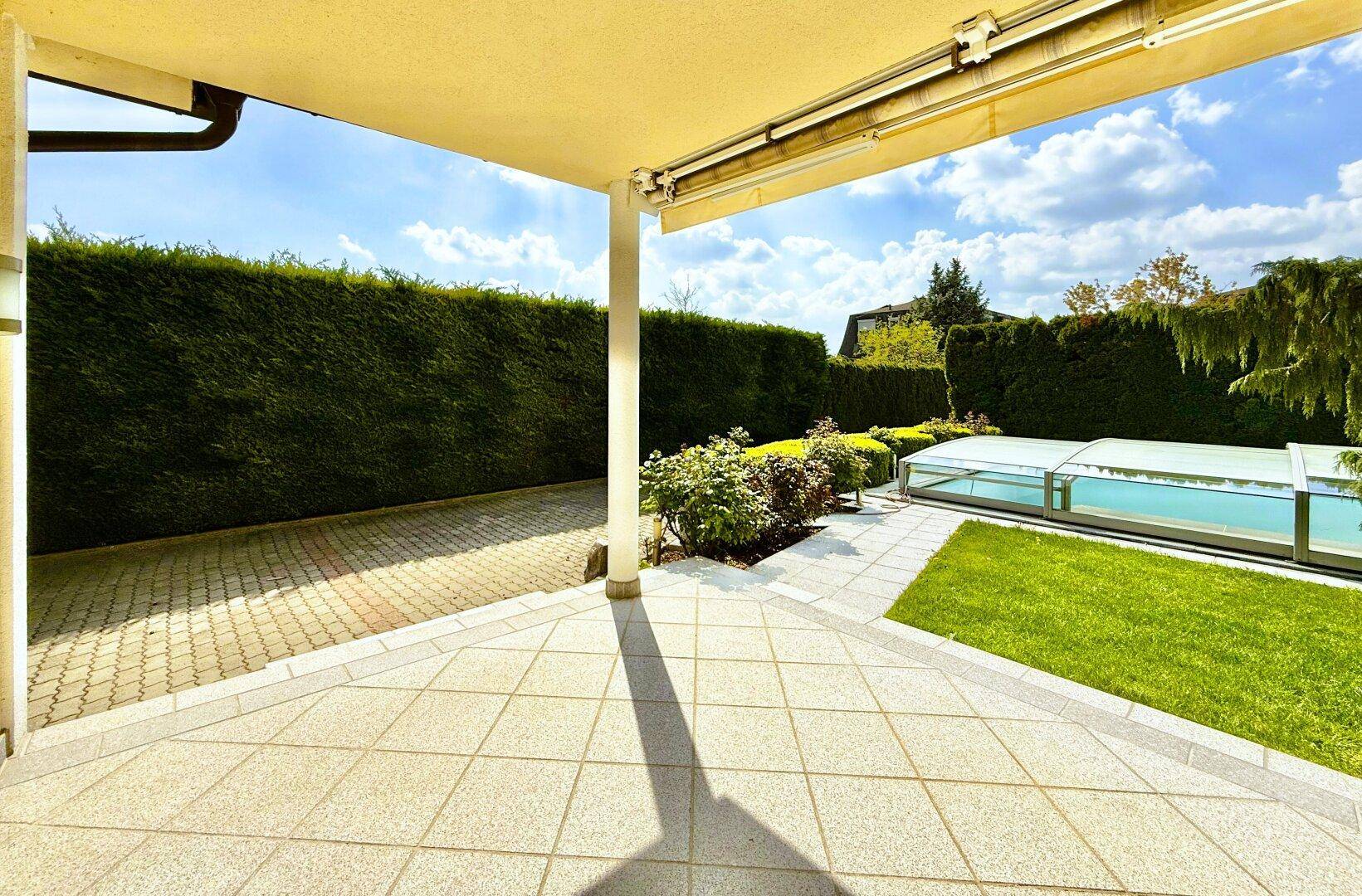 Überdachte Terrasse mit Fliesenboden und Blick auf den gepflegten Gartenbereich mit Pool.