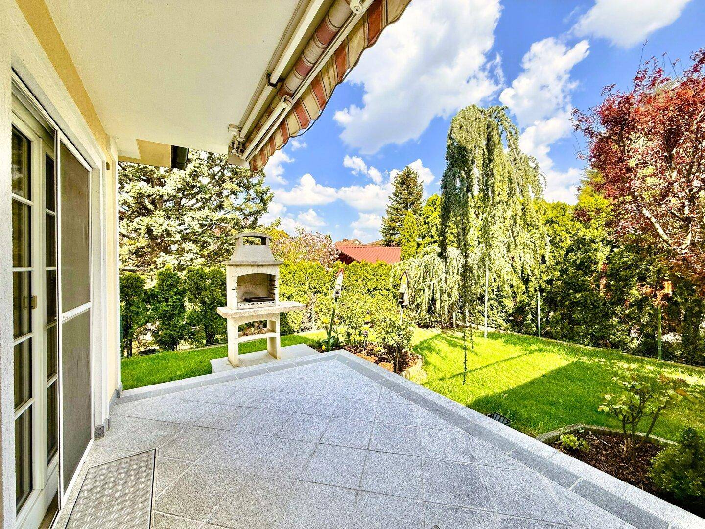 Geflieste Terrasse mit Außenkamin und Blick auf den grünen Gartenbereich unter blauem Himmel.
