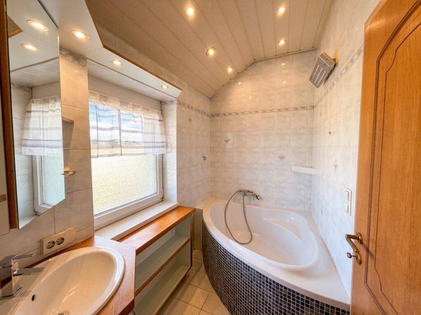 Badezimmer mit Eckbadewanne, Waschbecken, Fenster und Holzdecke in hellem Design.