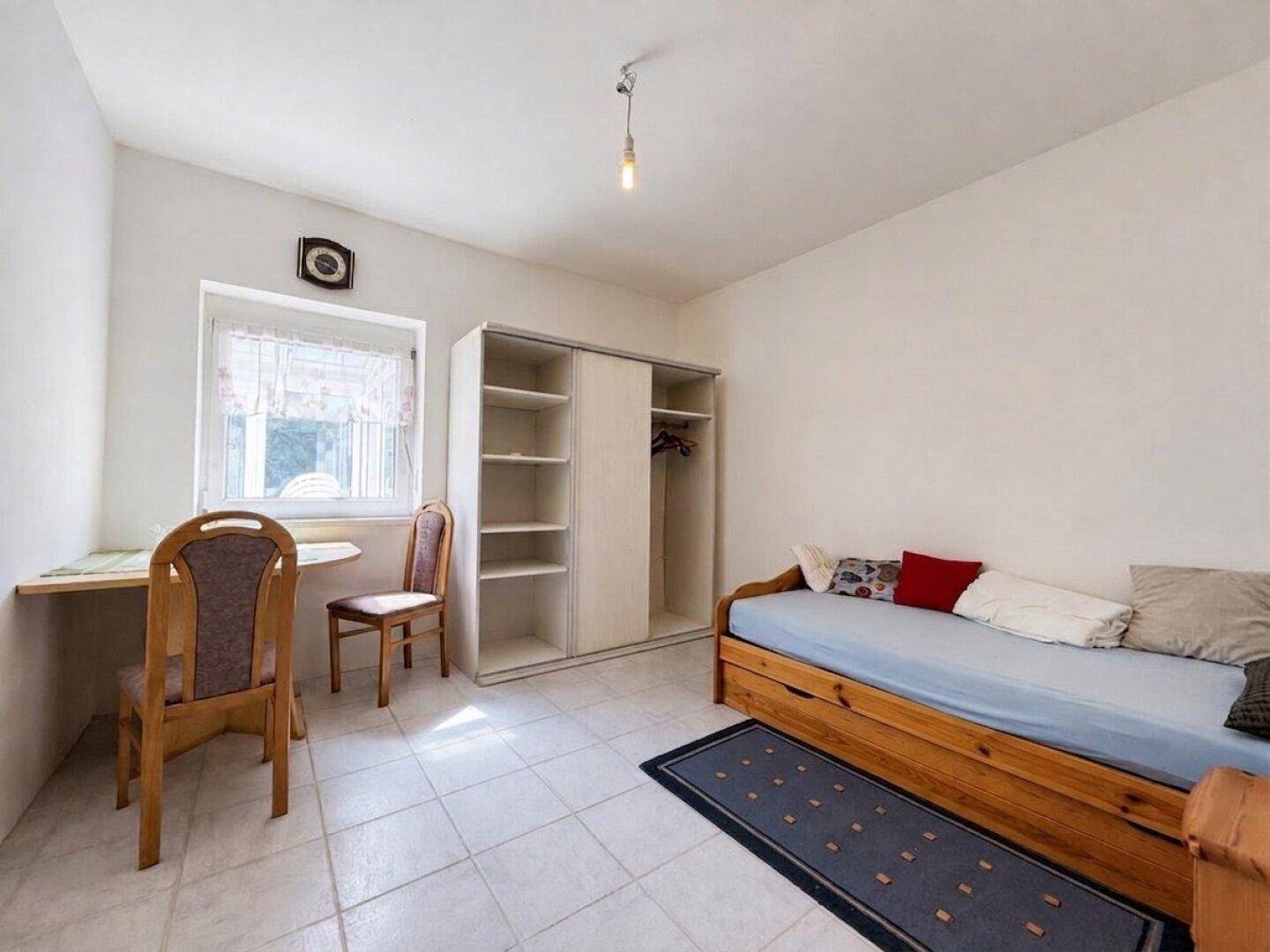 Helles Schlafzimmer mit Fliesenboden, Kleiderschrank und einem Arbeitsbereich am Fenster.