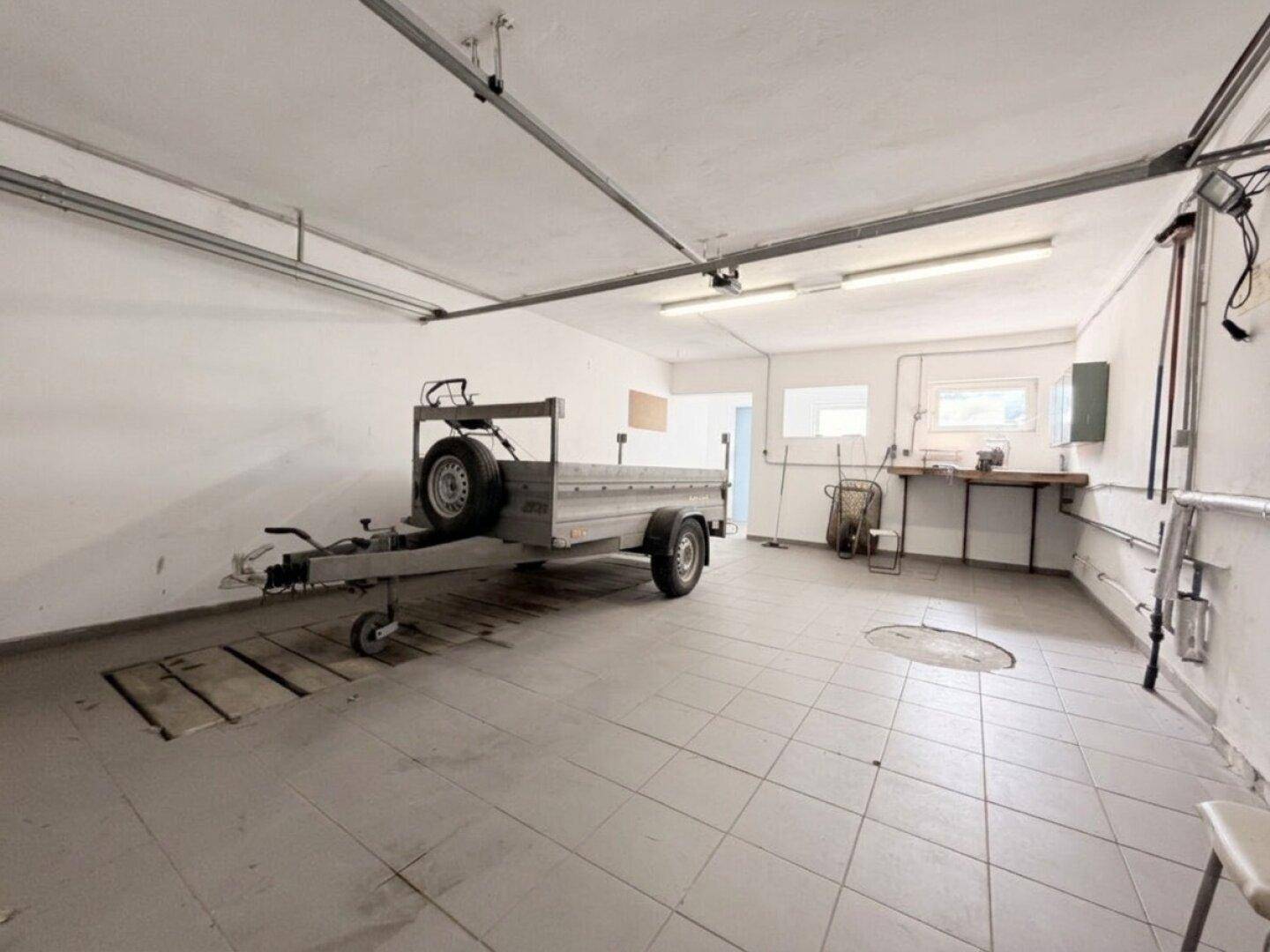 Geräumige Garage mit Fliesenboden, Werkbank und Platz für Fahrzeuge oder Anhänger.