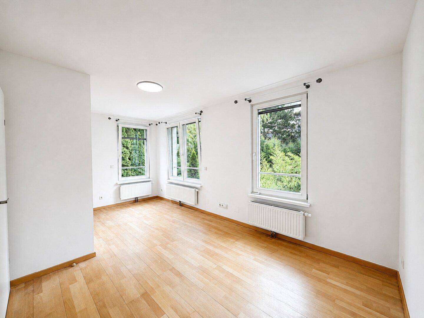 Helles Zimmer mit Holzfußboden und mehreren Fenstern, die viel Licht und grüne Aussicht bieten.