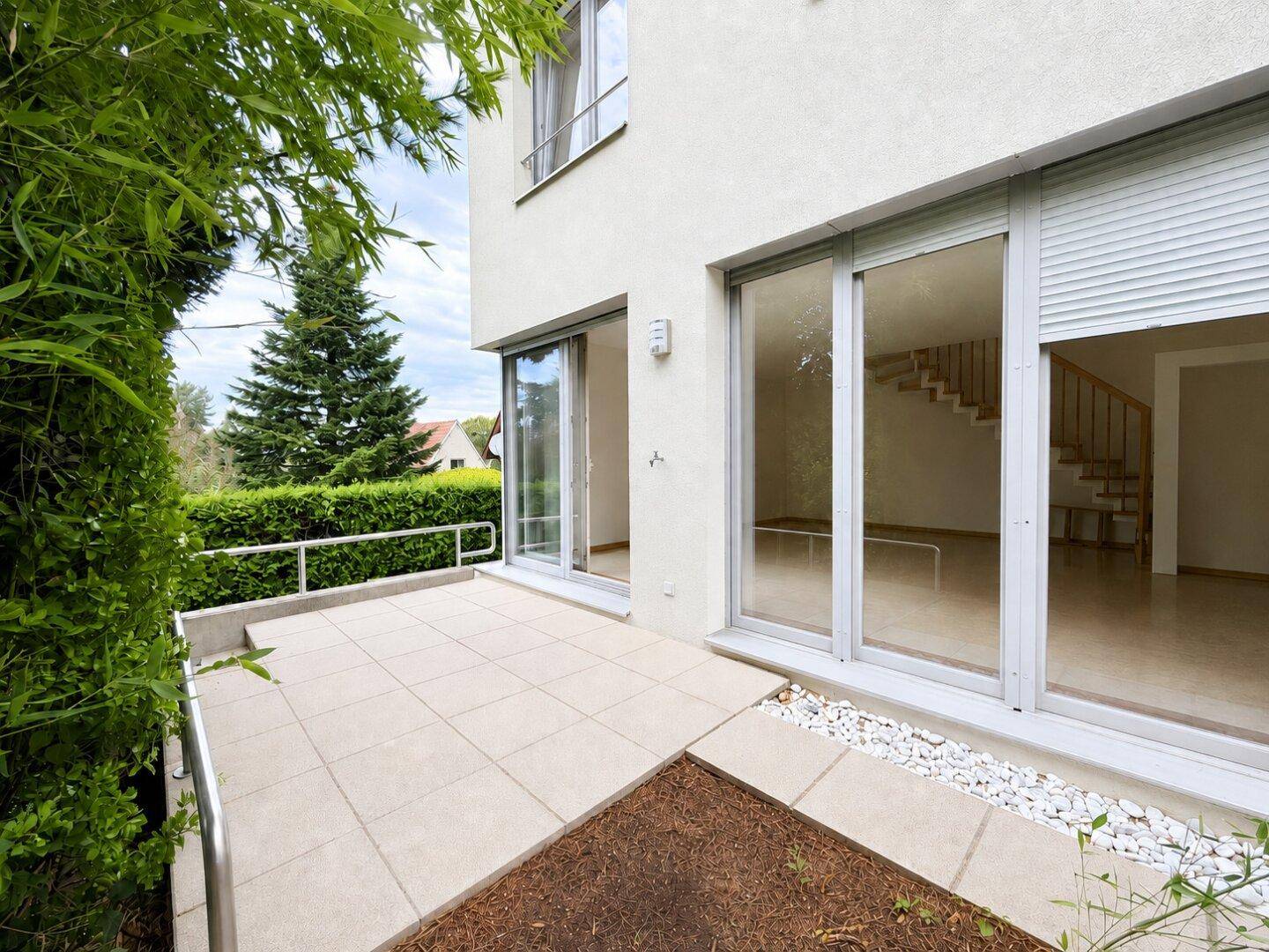 Leere Terrasse mit Fliesenboden und Zugang zum Innenbereich, umgeben von Grün.