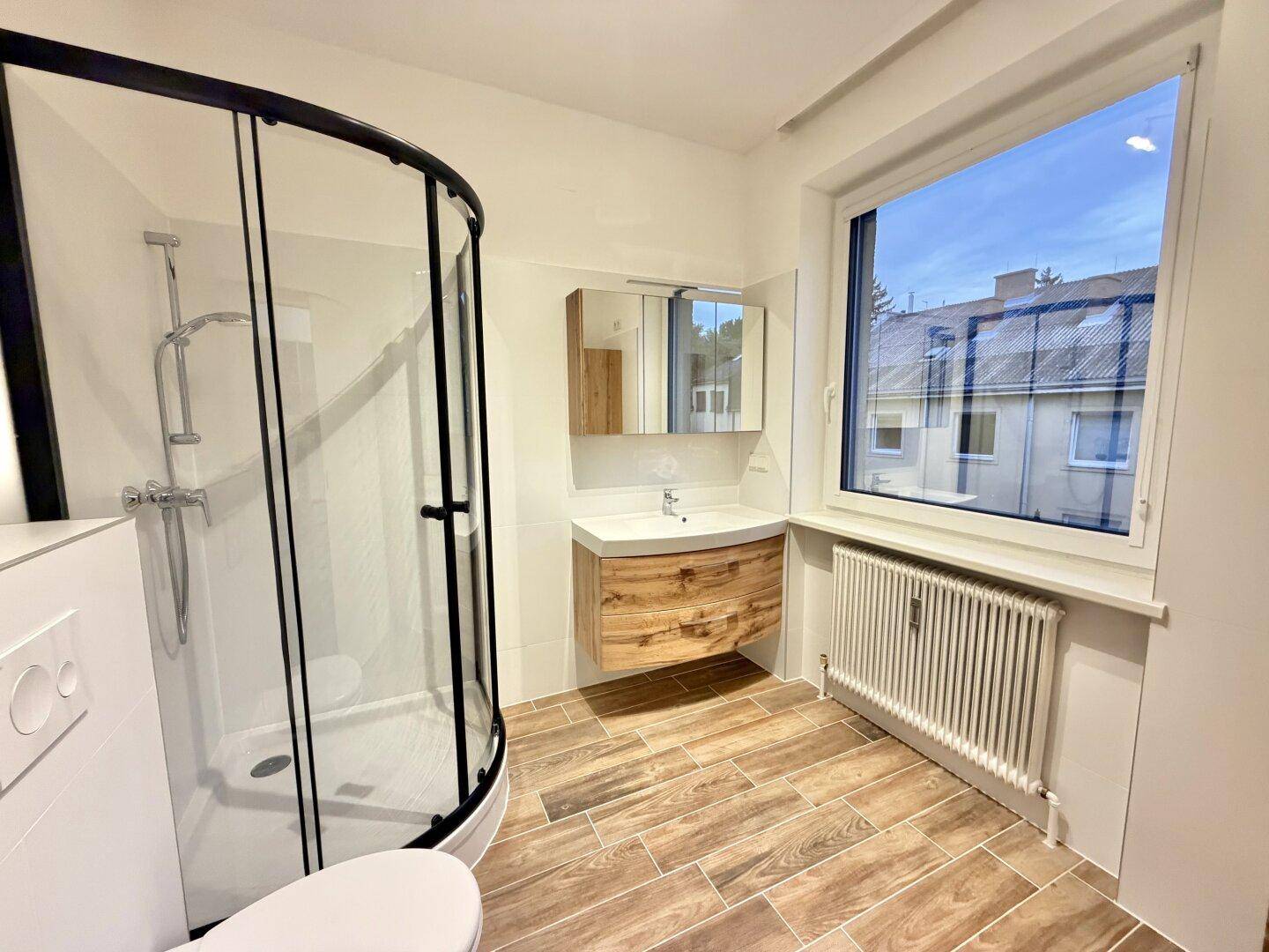 Modernes Badezimmer mit runder Glasdusche, Holzwaschtisch und einem Fenster.