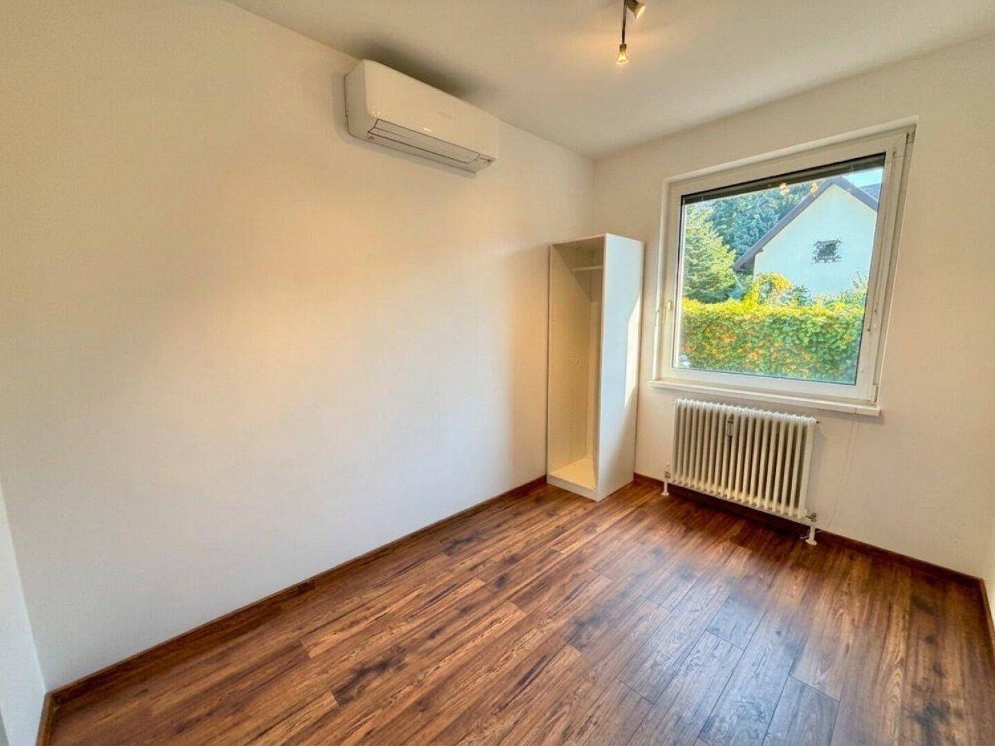 Schlafzimmer mit Holzfußboden, Klimaanlage und einem Fenster mit Blick ins Grüne.