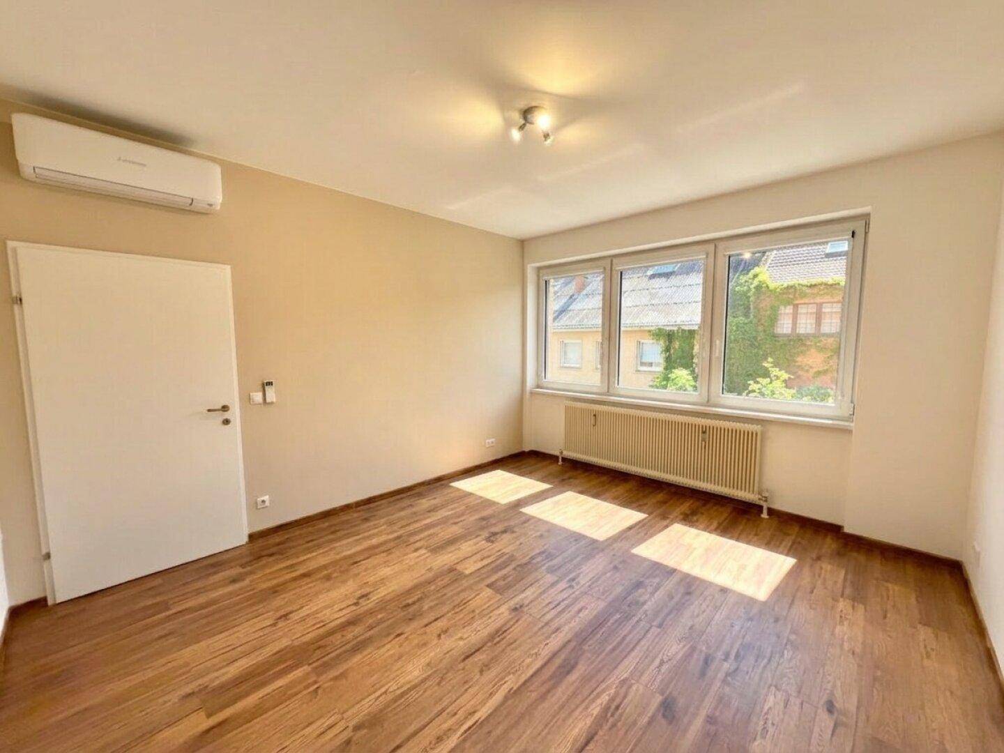 Geräumiger Raum mit Holzfußboden, Klimaanlage und großen Fenstern, die viel Licht hereinlassen.