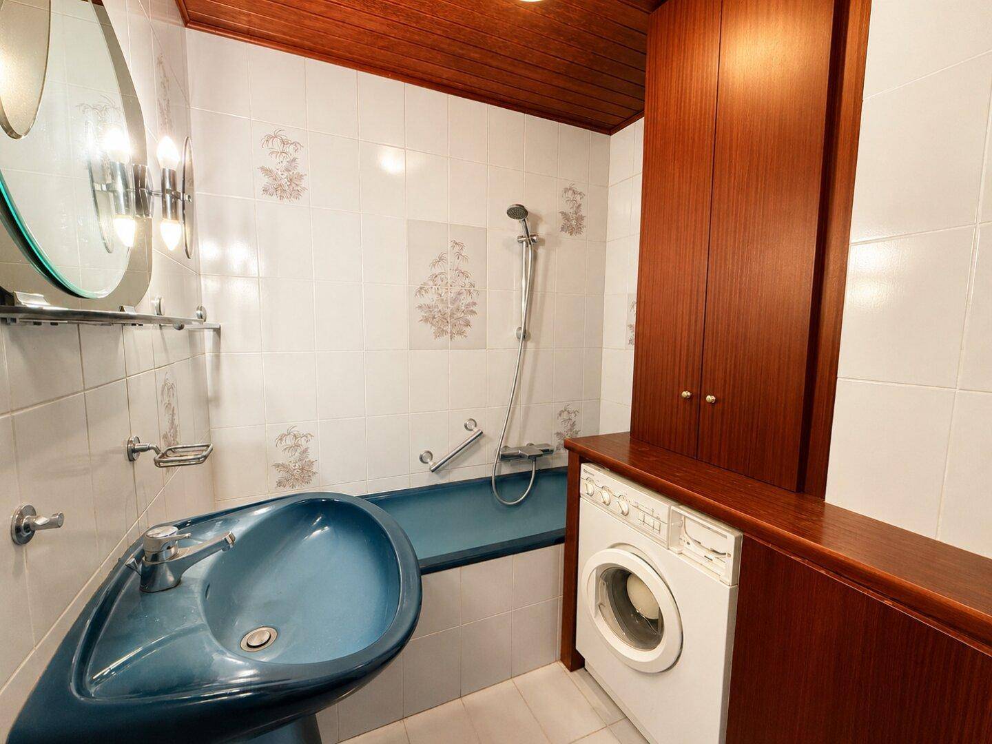 Badezimmer mit blauer Badewanne, Waschmaschine und Holzdecke im 80er-Jahre-Stil.