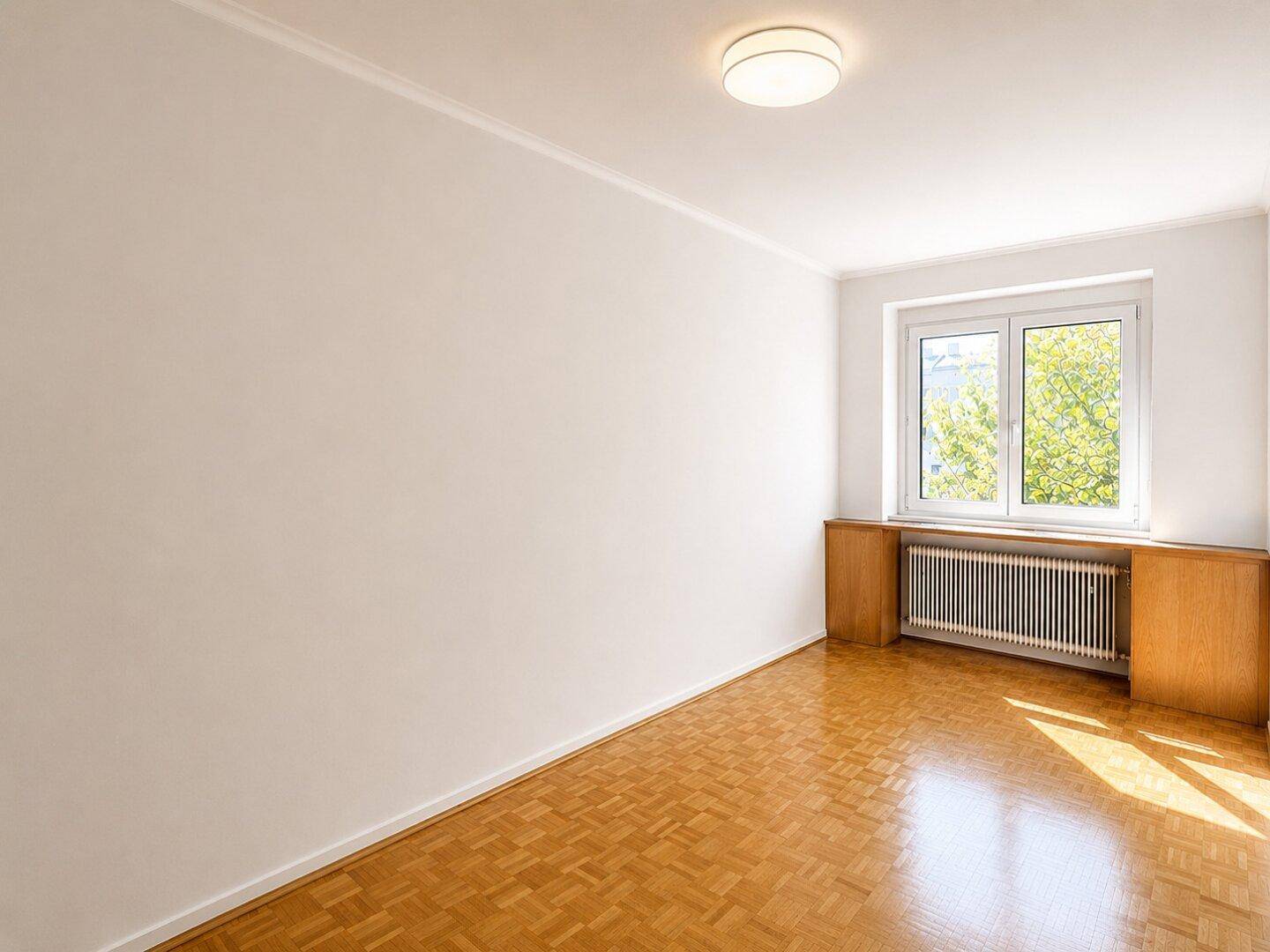 Helles, leeres Zimmer mit Parkettboden und Fenster mit Blick ins Grüne.