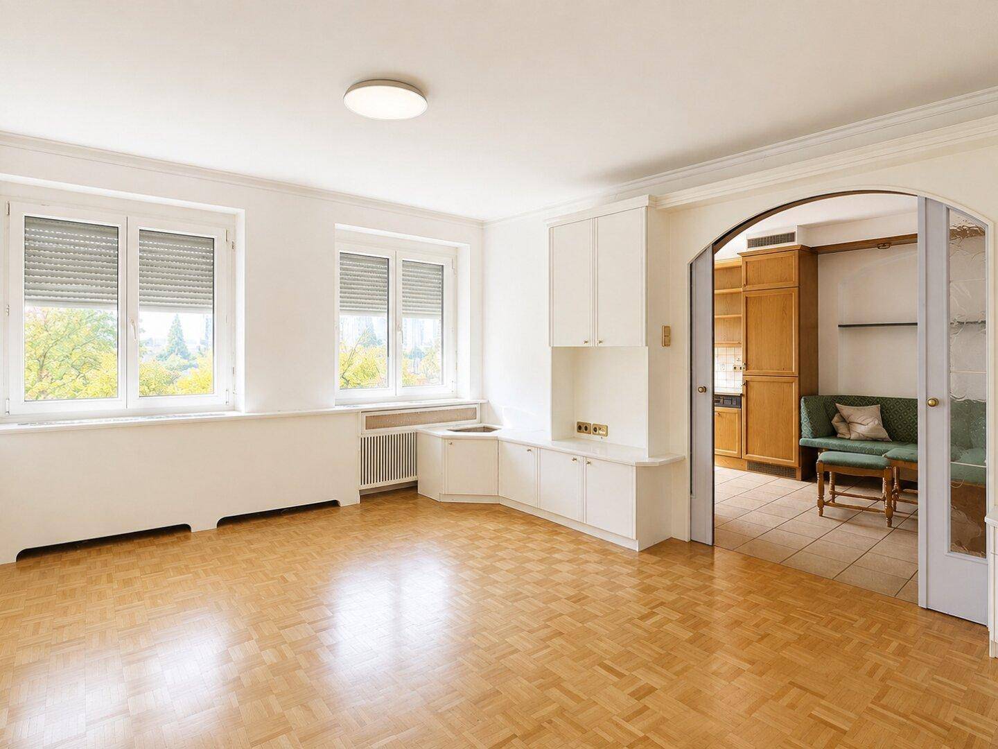 Helles, leeres Wohnzimmer mit Parkettboden, Rollläden und Durchgang zur Küche.