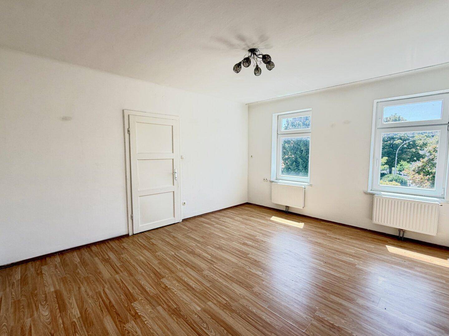 Leerer Raum mit Holzimitat-Laminatboden, weißen Wänden und zwei Fenstern, die viel Licht hereinlassen.
