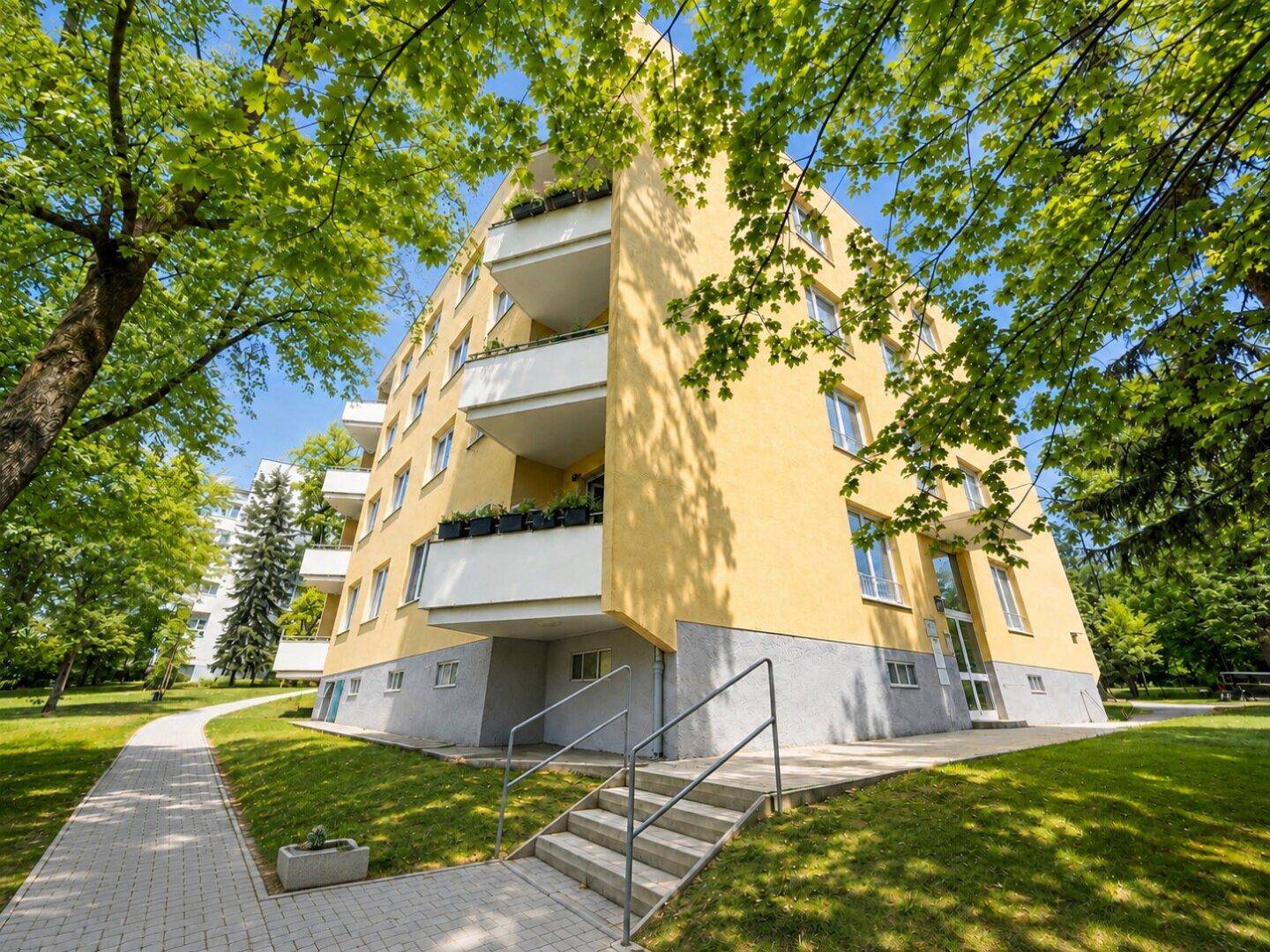 Gelbes Mehrfamilienhaus mit Balkonen, umgeben von Bäumen und einer Grünanlage.