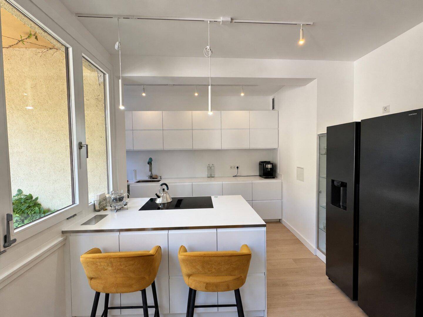 Moderne weiße Küche mit Kochinsel, Induktionskochfeld und schwarzen Kühlgeräten, sehr funktional.