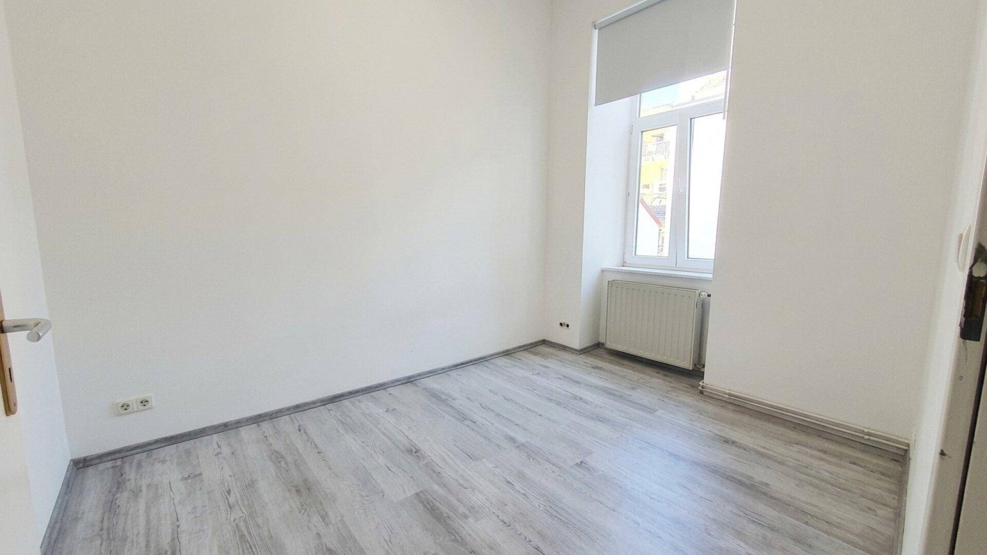Leerer Raum mit hellem Laminatboden und einem Fenster mit Rollo für Privatsphäre.