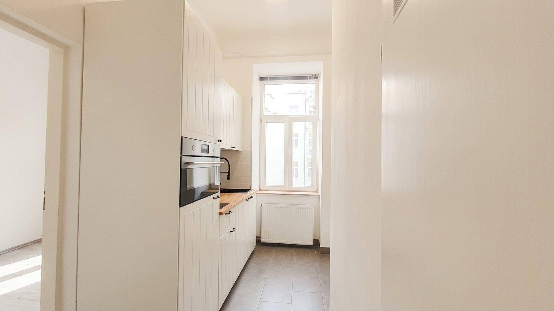 Helle Küche mit weißen Schränken, Einbauofen, Holzarbeitsplatte und Fenster für Tageslicht.