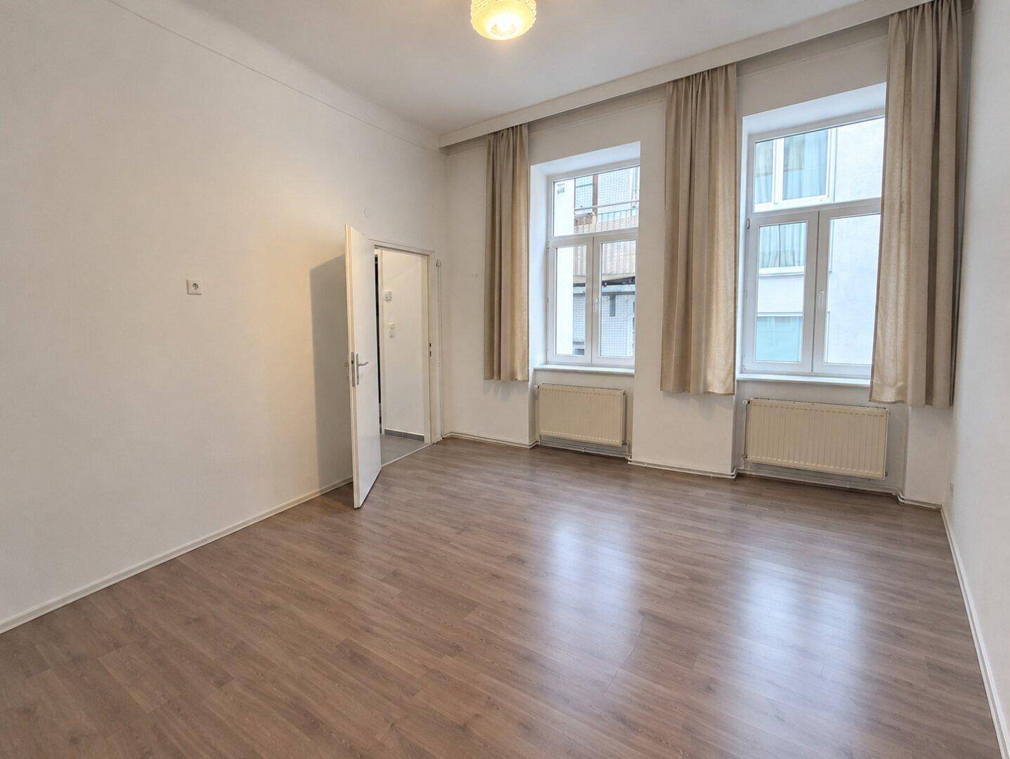 Leerer Raum mit Laminatboden, hohen Decken und zwei großen Fenstern, bereit zur Einrichtung.