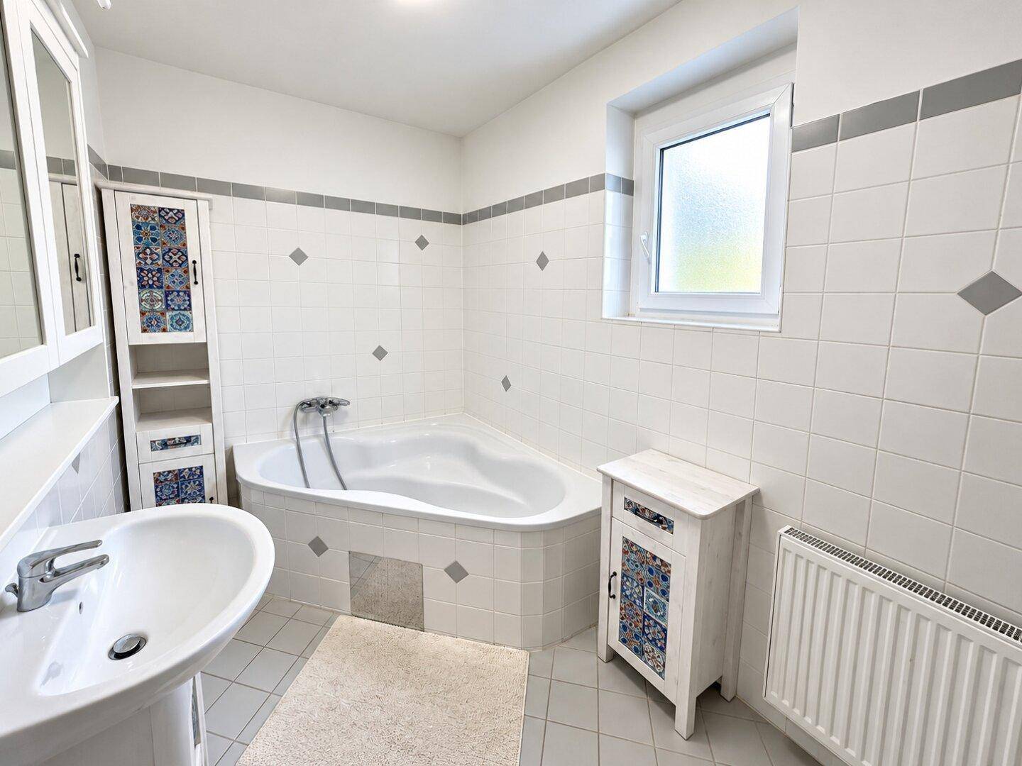Helles Badezimmer mit weißen Fliesen, einer Eckbadewanne, Waschbecken und Fenster.