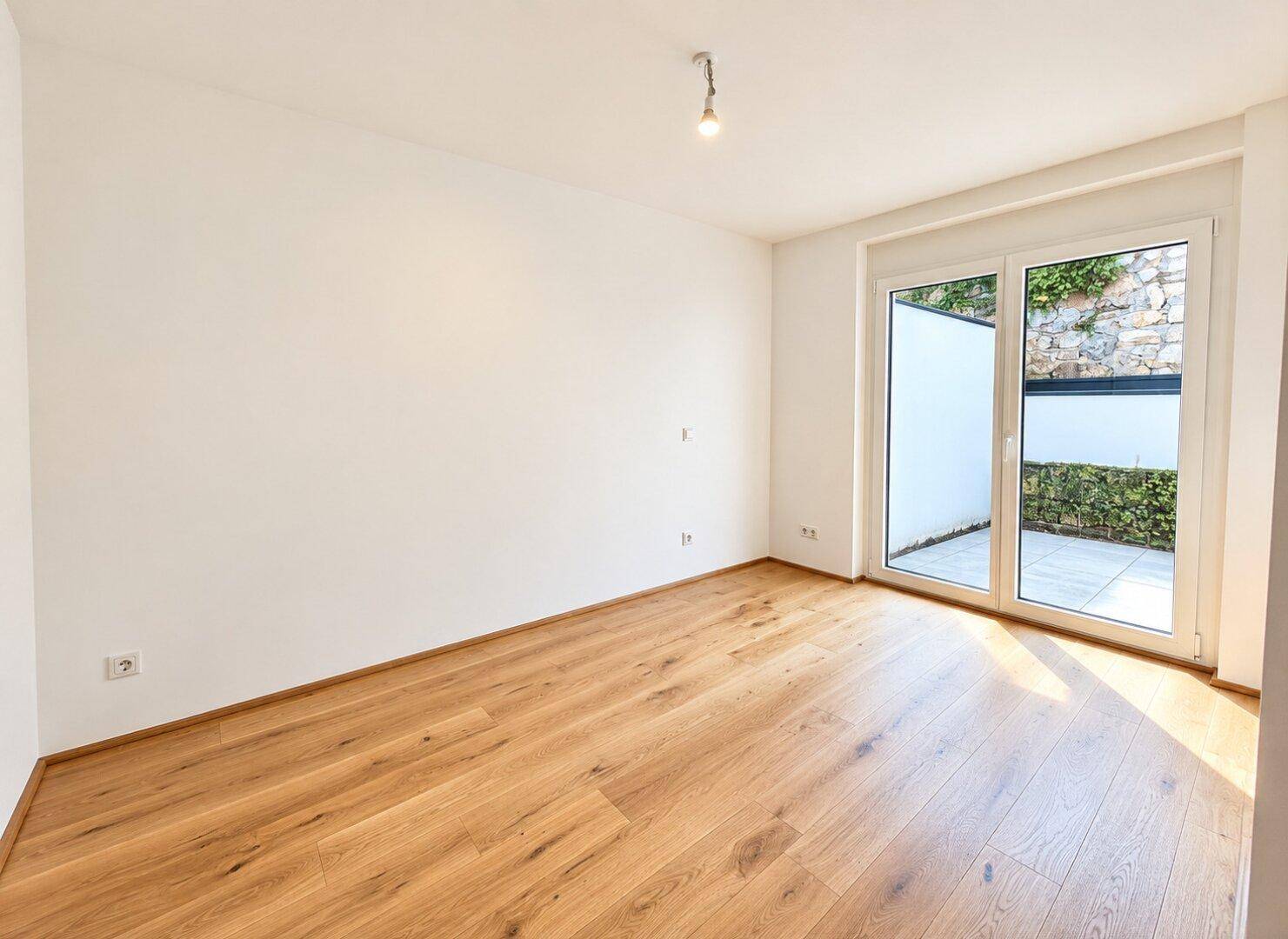 Heller Raum mit Holzboden und großem Fenster, das Zugang zu einem kleinen Außenbereich bietet.
