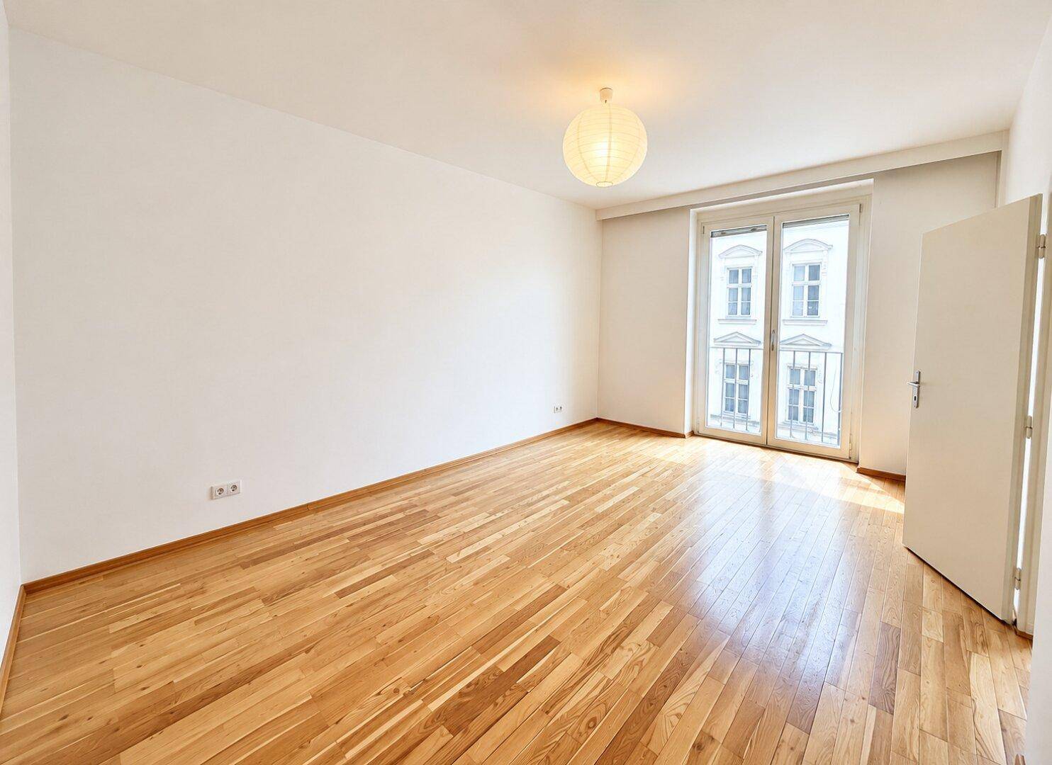 Geräumiges Schlafzimmer mit hellem Holzboden, weißen Wänden und einem französischen Balkon.