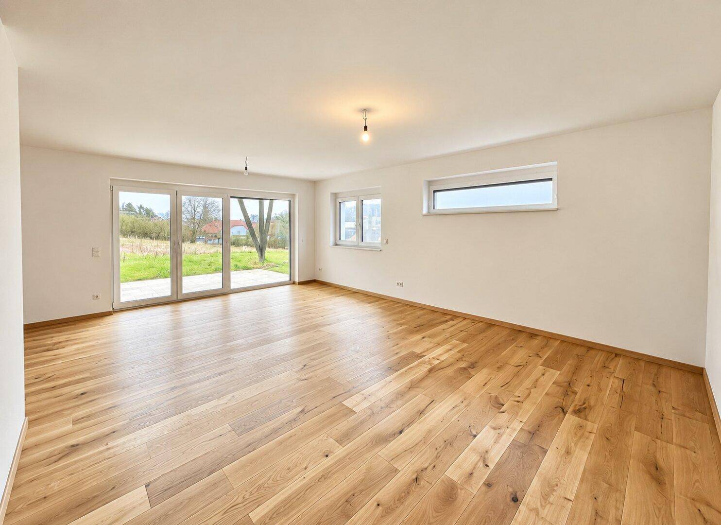 Geräumiges, leeres Wohnzimmer mit hellem Holzboden und großen Fenstern zum Garten.