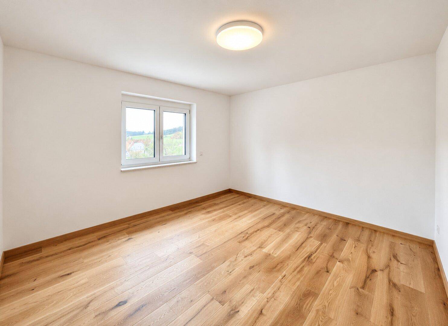 Helles, leeres Zimmer mit Holzboden und Fenster, ideal als Kinder- oder Gästezimmer.