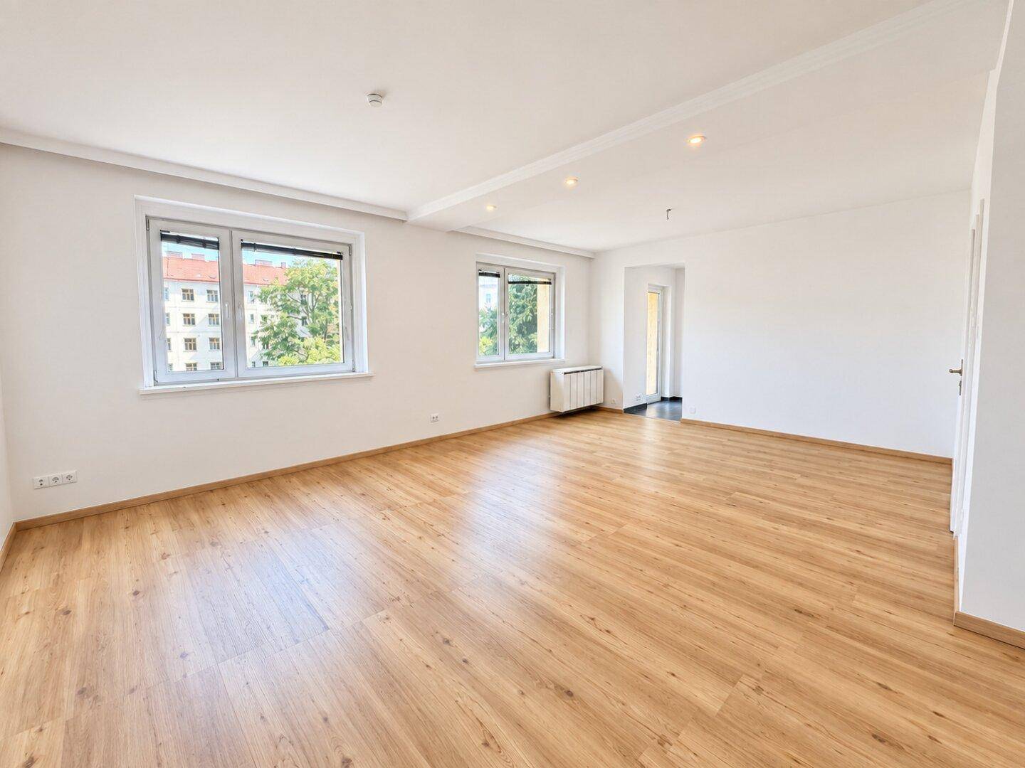 Heller, leerer Wohnraum mit Laminatboden und großen Fenstern, die viel Tageslicht hereinlassen.