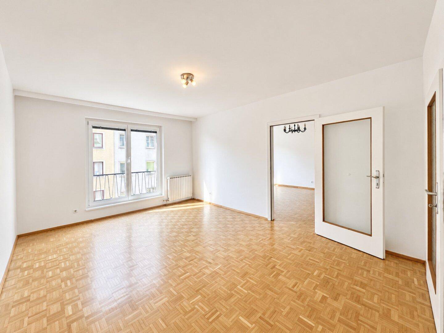 Helles, leeres Wohnzimmer mit Fischgrätparkett, weißen Wänden und französischem Balkon.
